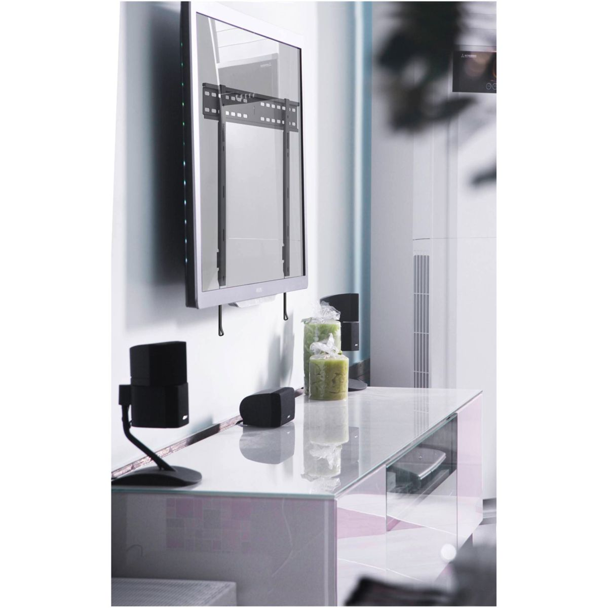 Fernseher an der Wand montiert, mit Glasregal darunter. Lautsprecher, Dekorationen und ein weißer Schrank sind sichtbar.