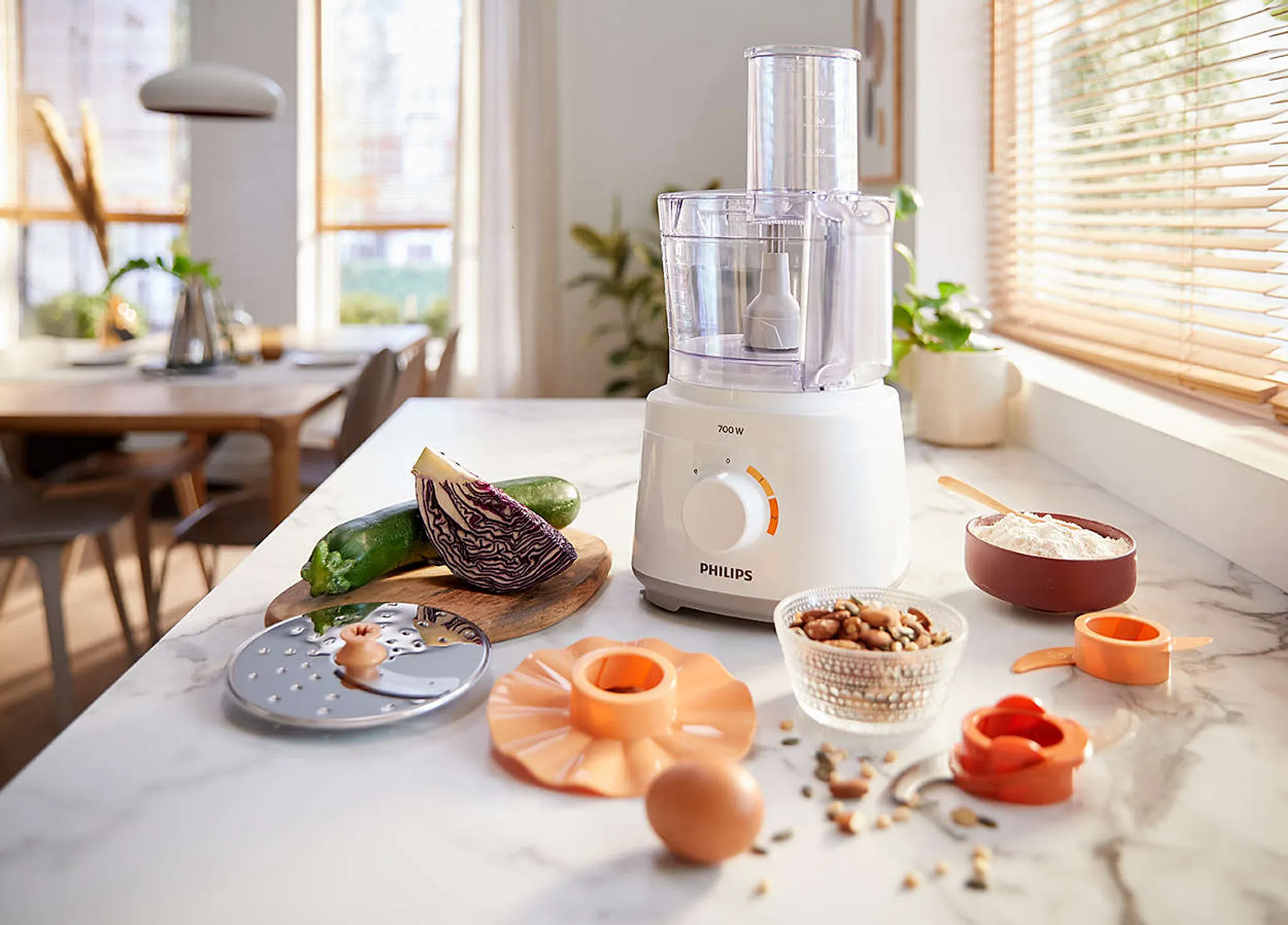 A white and clear food processor with vegetables, nuts, flour, and other ingredients on a counter.
