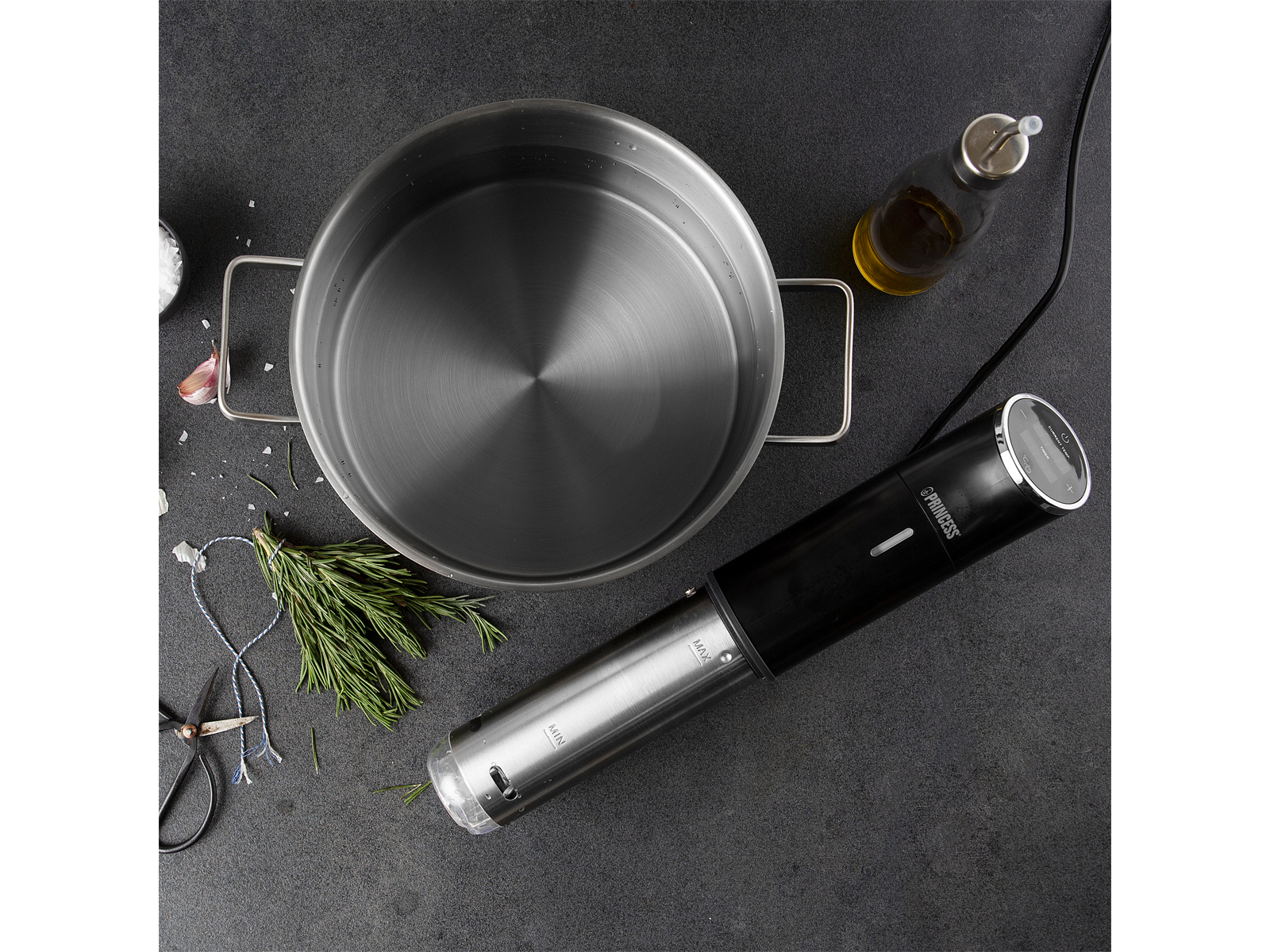 Una olla de acero inoxidable y una máquina de sous vide en una superficie oscura. Romero y aceite cerca.
