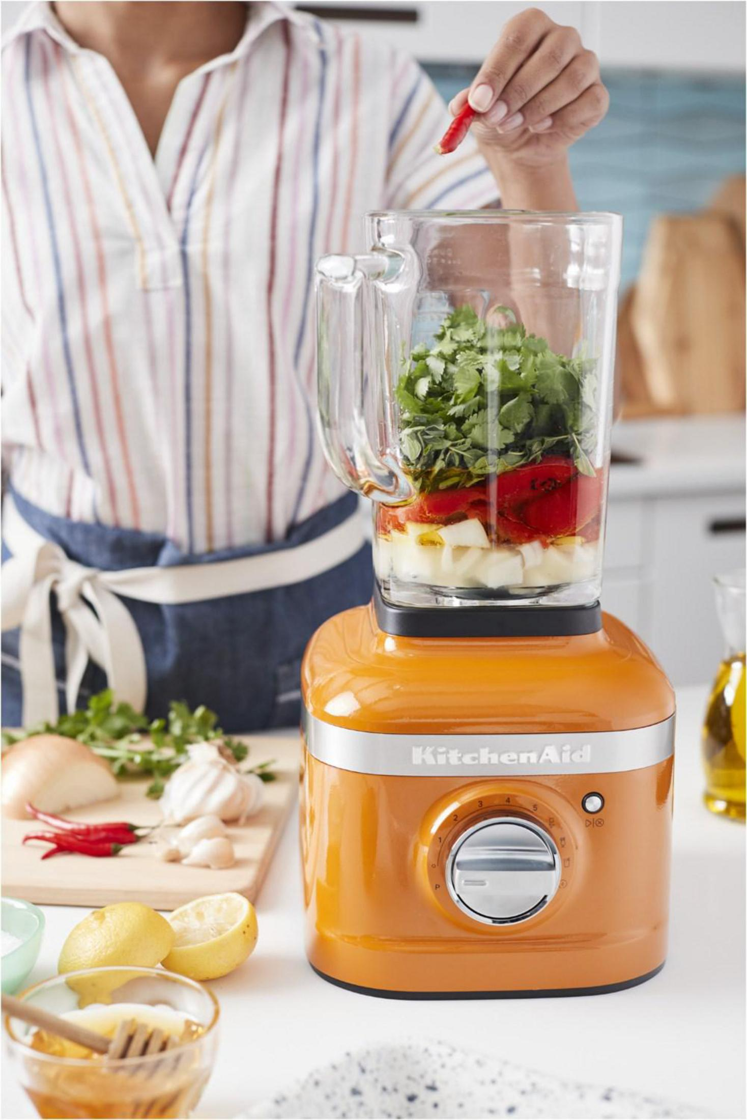 A person making a sauce in an orange KitchenAid blender, with ingredients visible inside.