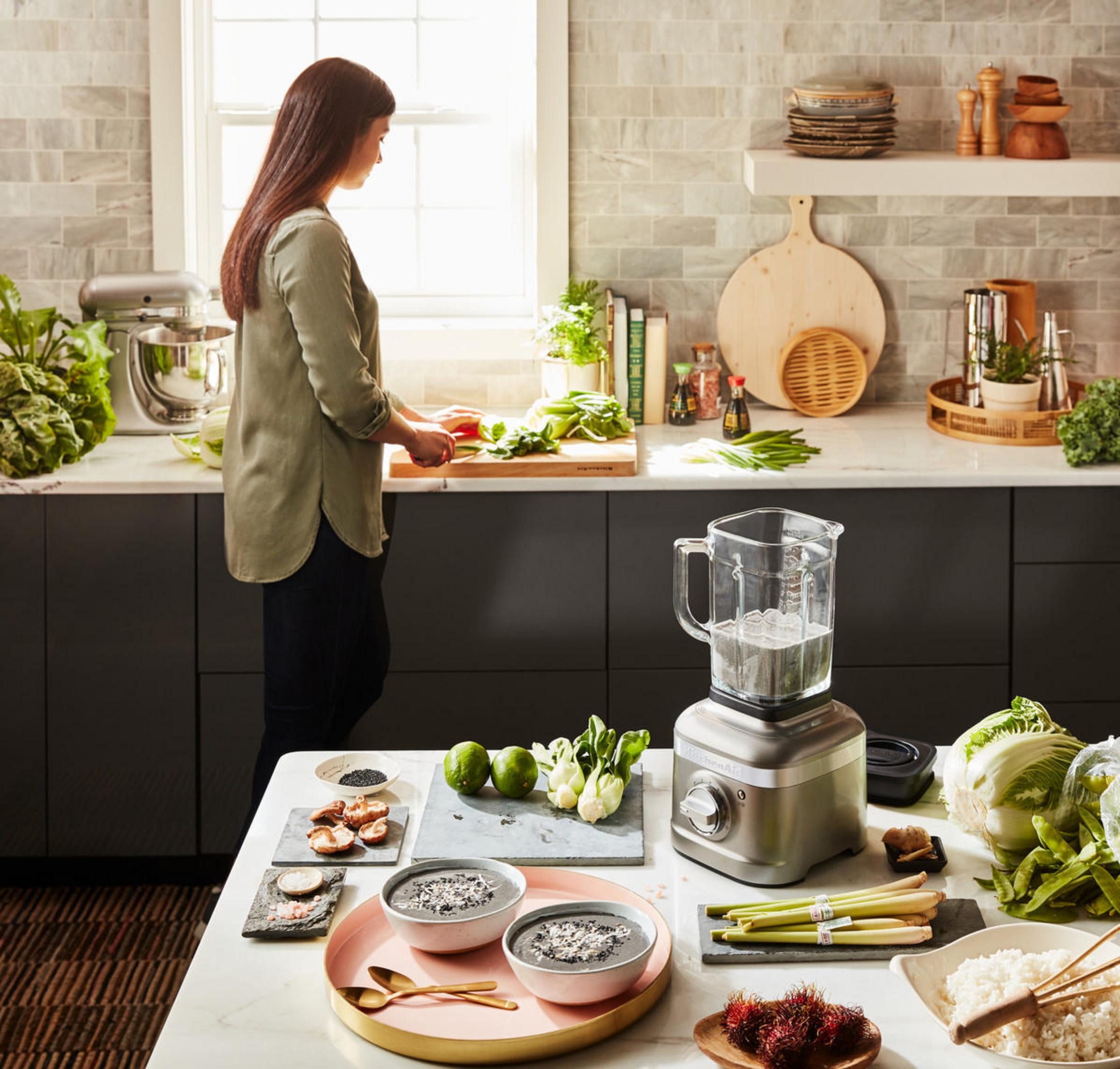 Kobieta kroi warzywa w kuchni. Blender, składniki i miski na blacie. Jasna, słoneczna scena.