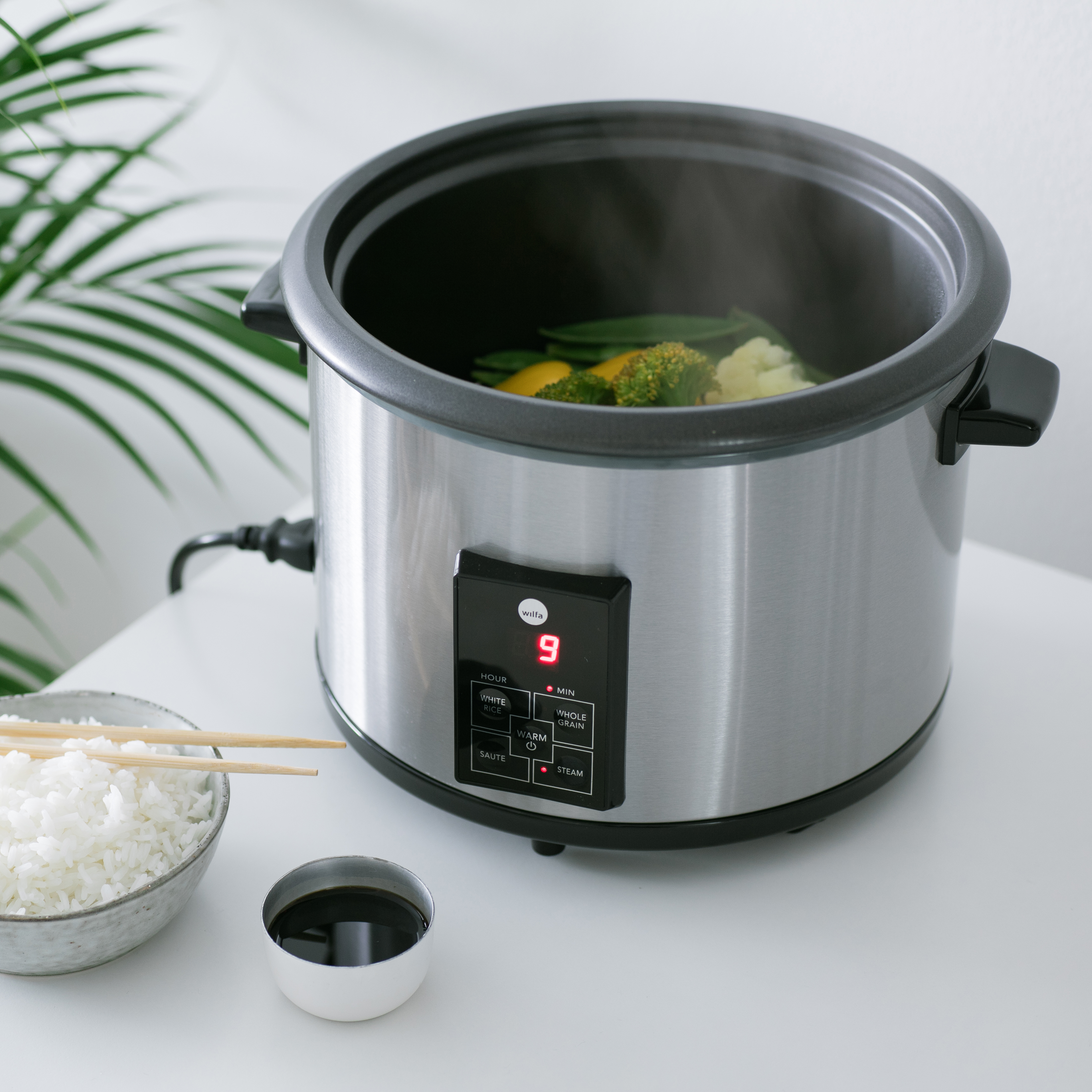 Ein silberner Slow Cooker mit Gemüse, Essstäbchen und einer Schüssel mit Reis und Soße.