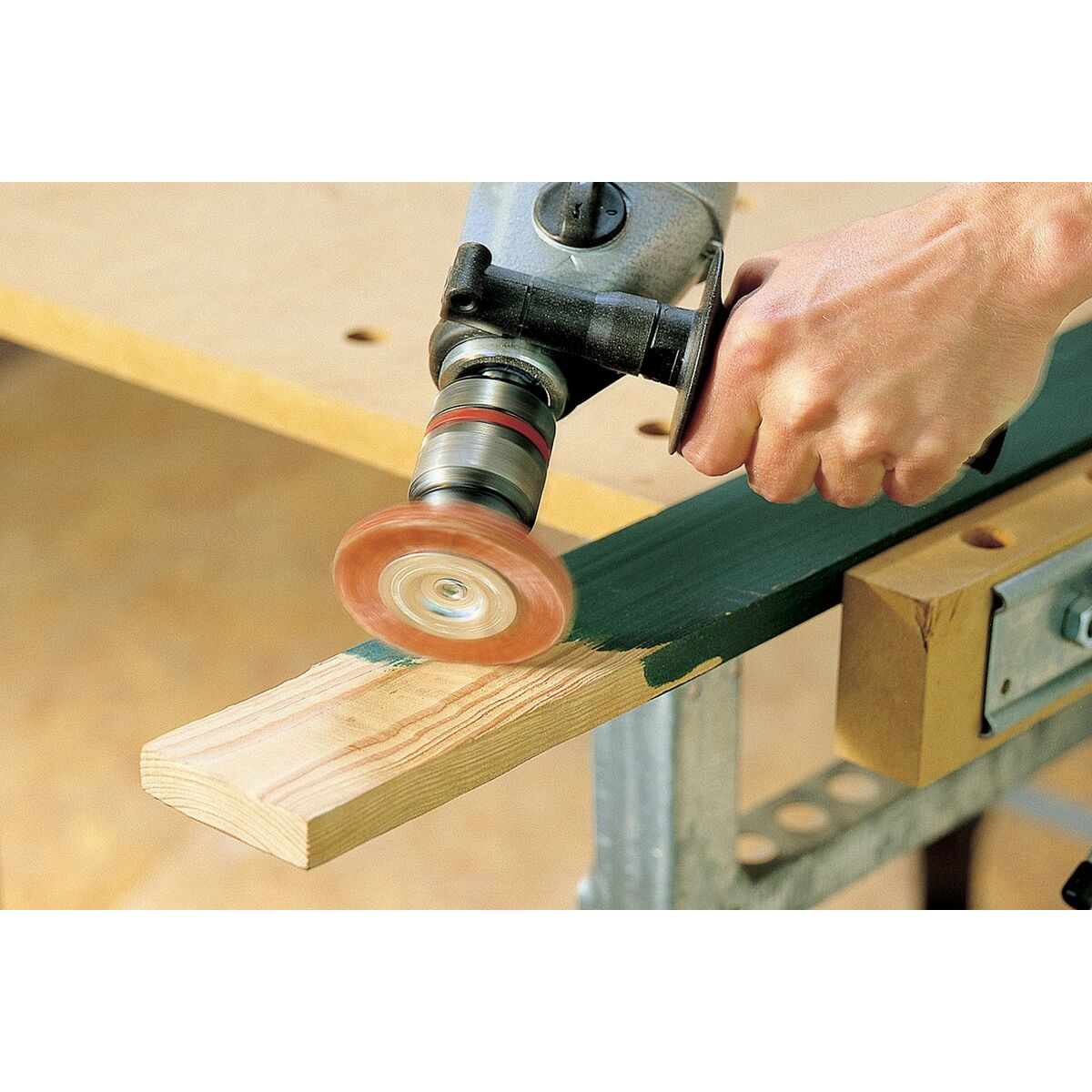 Una persona lijando una tabla de madera pintada de verde con una herramienta eléctrica y una almohadilla de lijado naranja.