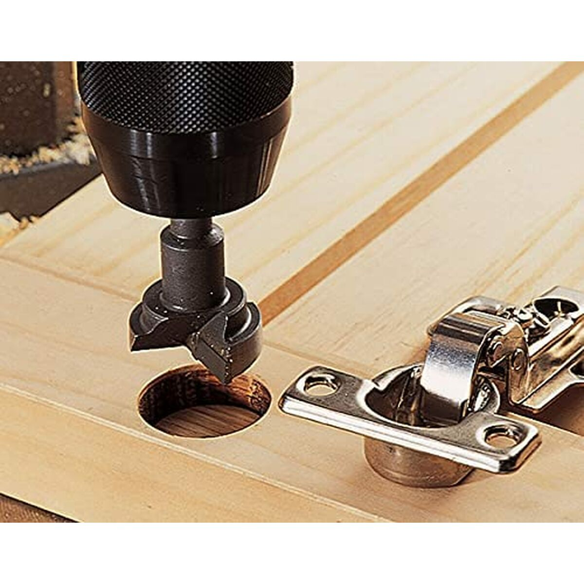 Una broca taladrando un agujero en madera. Una bisagra está cerca. La escena está sobre una superficie de madera.