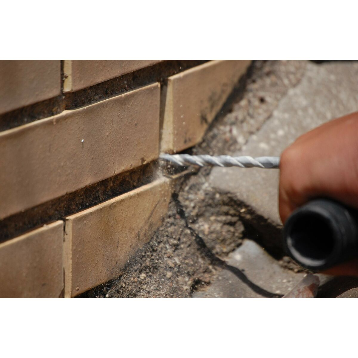 Una mano taladra una pared de ladrillos. El taladro es plateado y la pared es de color canela.