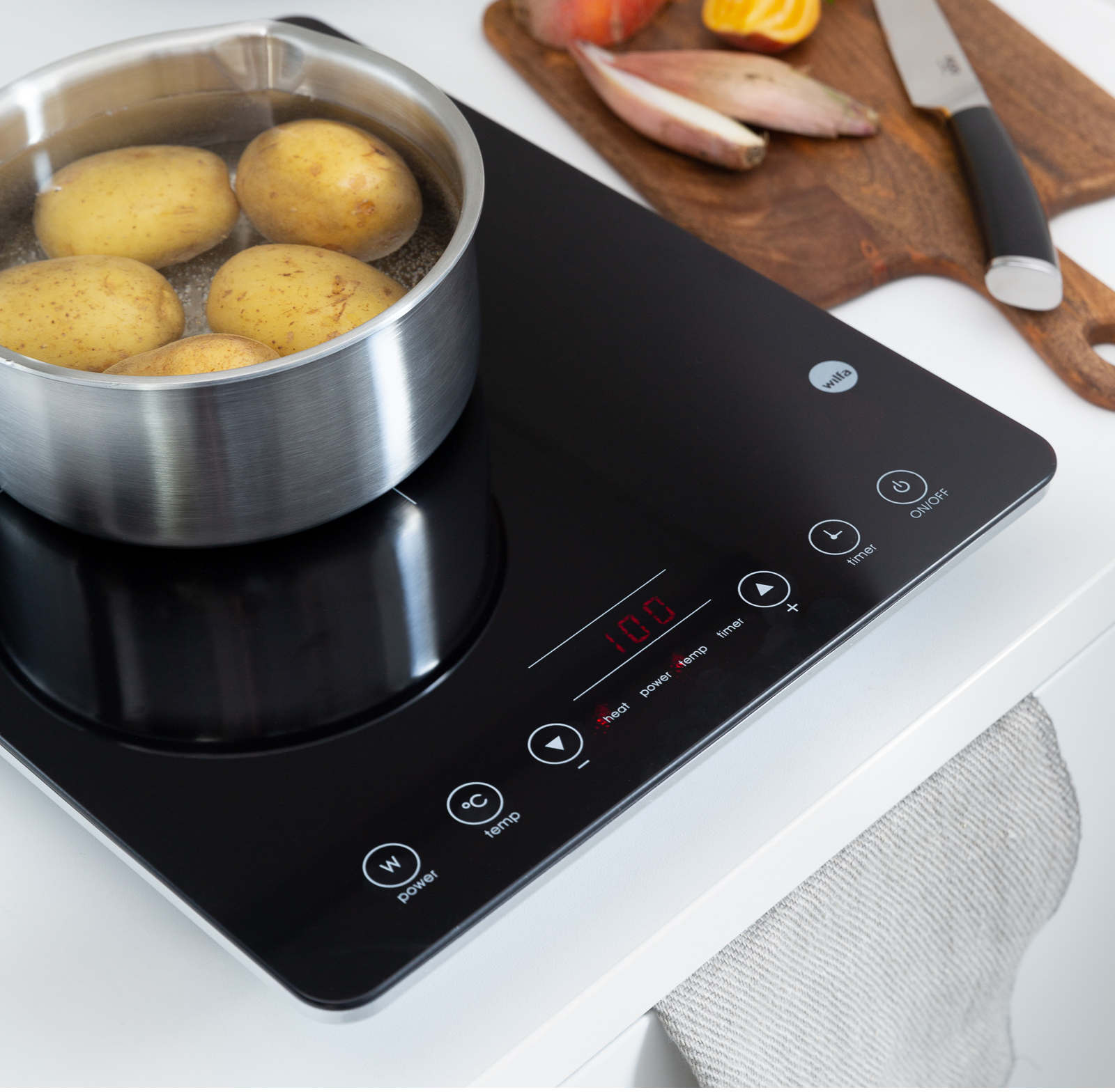 Placa de inducción negra con verduras y patatas en una olla, sobre superficie blanca.