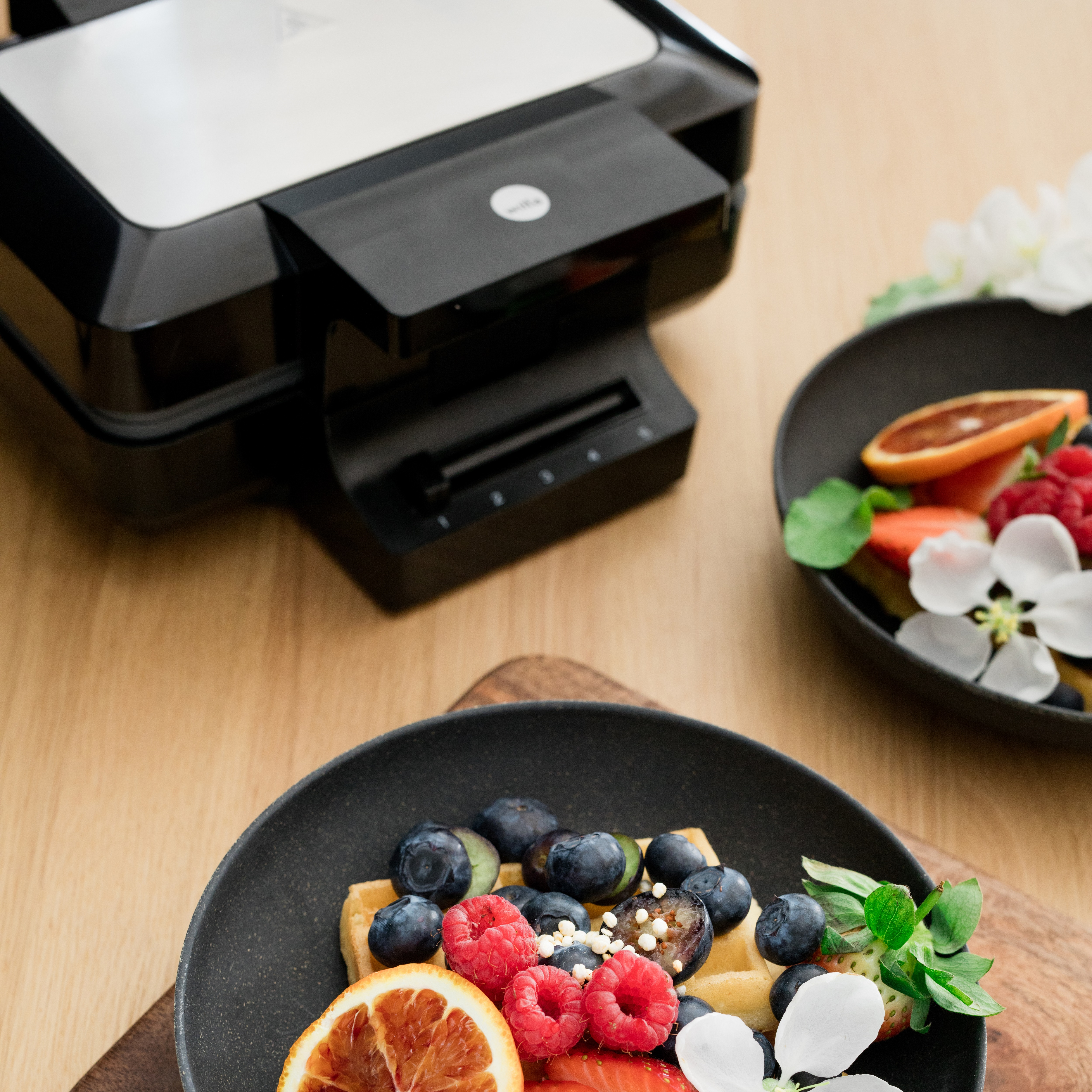 Waffeln mit Beeren auf einem Holzbrett. Ein Waffeleisen und ein Teller sind im Hintergrund.