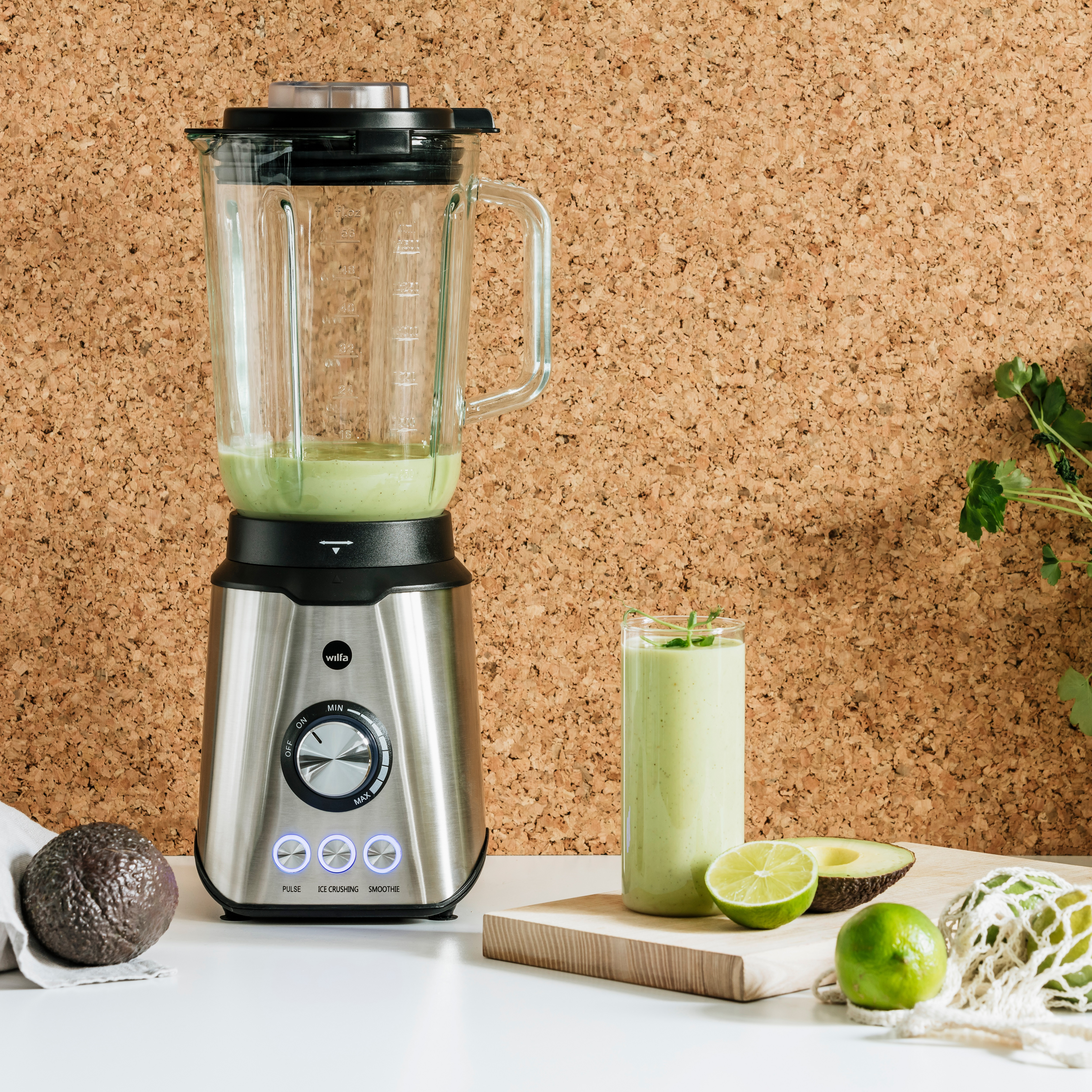 Mixer mit grünem Smoothie, Avocado, Limetten und Zutaten auf einer weißen Oberfläche, in der Nähe einer Pinnwand.