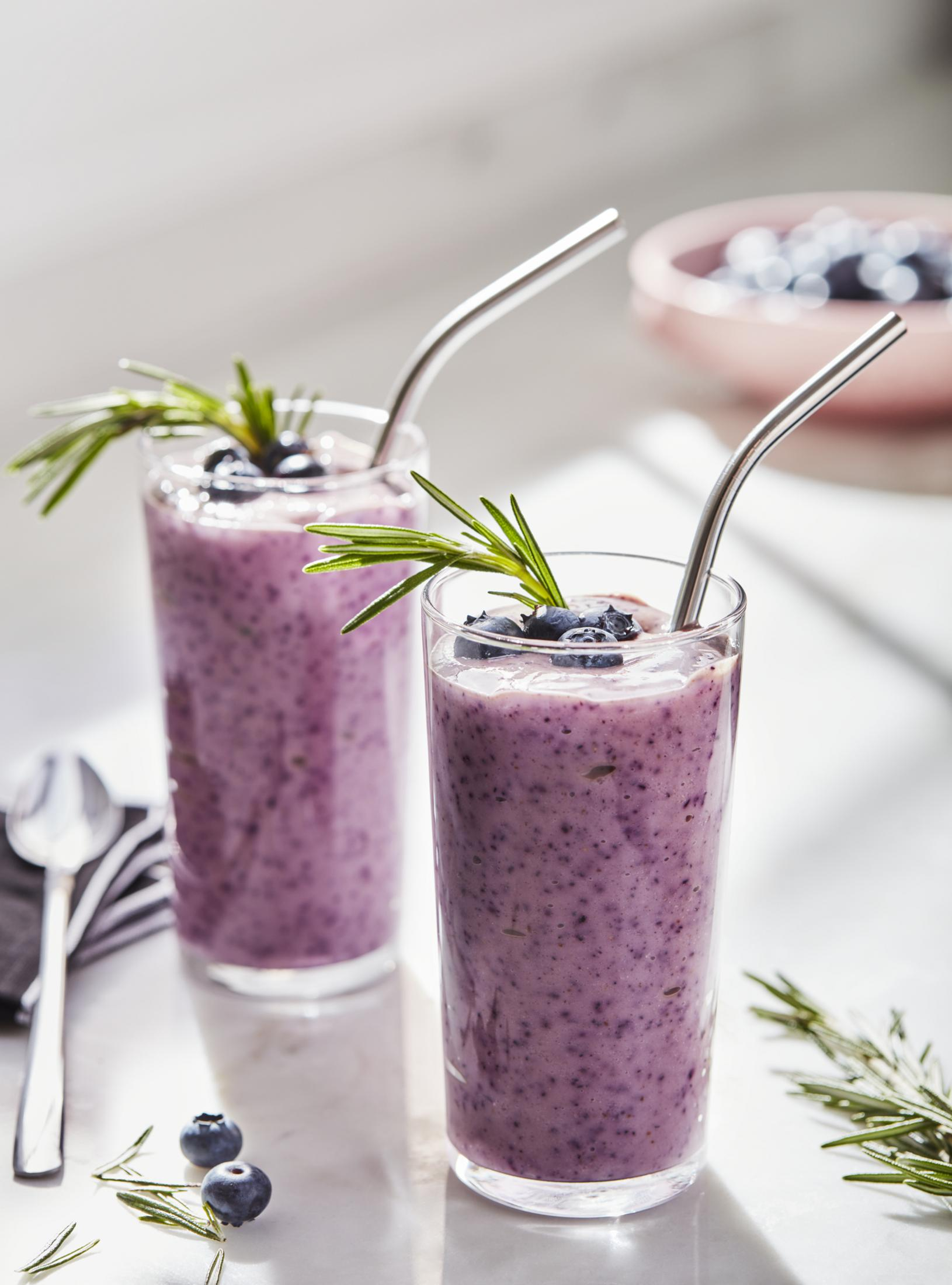 Zwei Blaubeer-Smoothies in hohen Gläsern mit Metallstrohhalmen, Rosmaringarnitur. Rosa Schüssel.