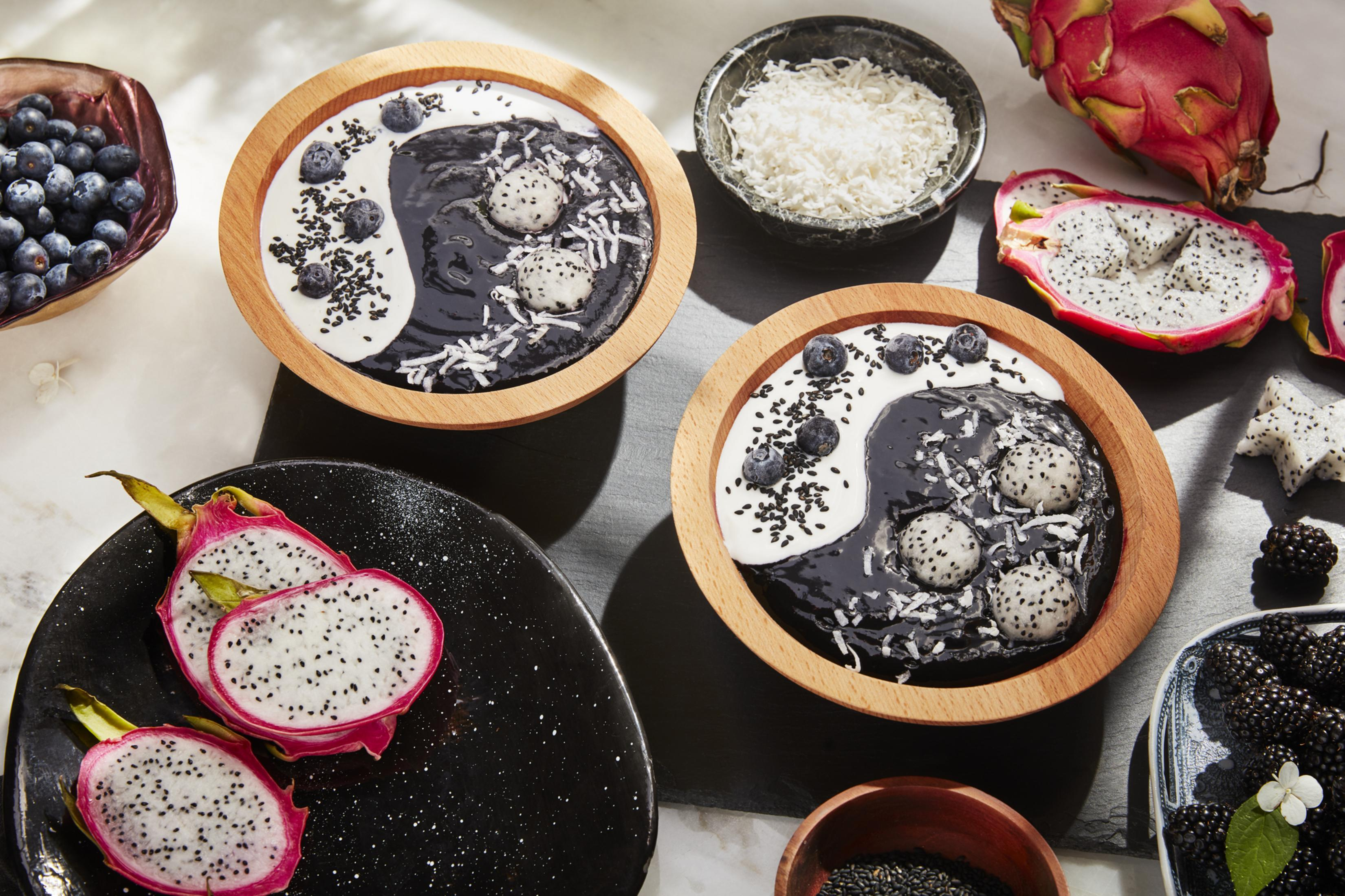 Zwei Holzschalen mit Yin-Yang-Smoothies mit Drachenfrucht, Blaubeeren und Kokosflocken.
