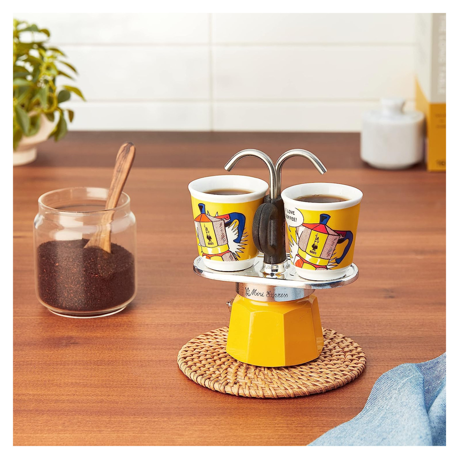 Żółta kawiarka z dwiema filiżankami kawy na drewnianym stole. Widoczny jest również słoik z ziarnami kawy i łyżka.