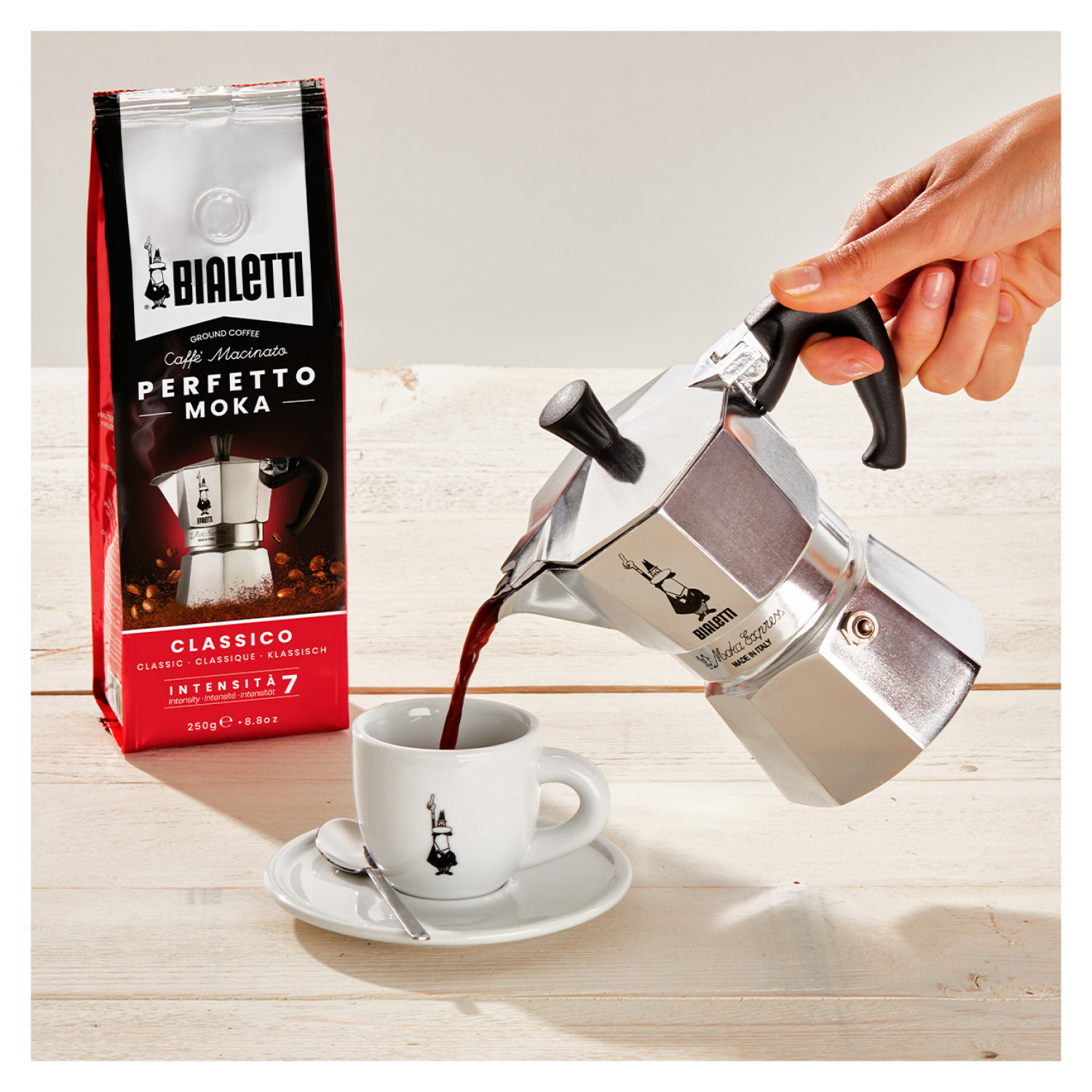 Osoba nalewa kawę ze srebrnego ekspresu Moka do białej filiżanki. Opakowanie kawy po lewej.