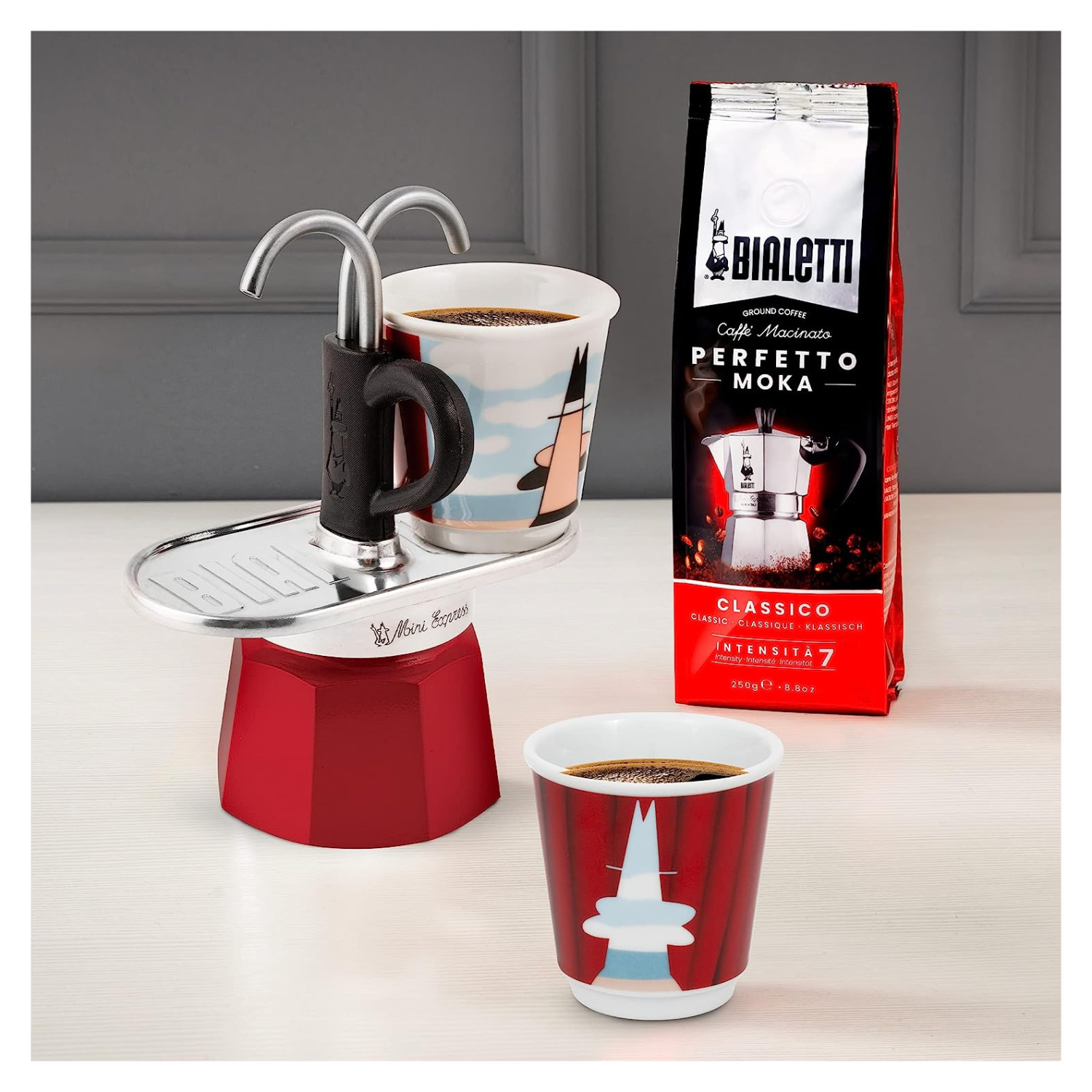 Czerwony ekspres do kawy Bialetti z dwiema filiżankami kawy i torebką ziaren kawy na białym stole.