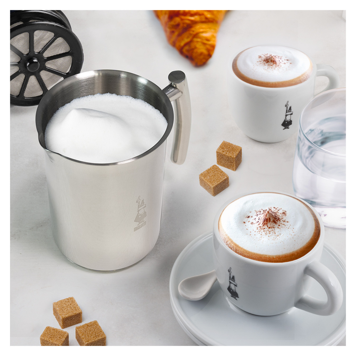 Spieniacz do mleka ze stali nierdzewnej z pianką, dwa cappuccino, kostki cukru i rogalik.