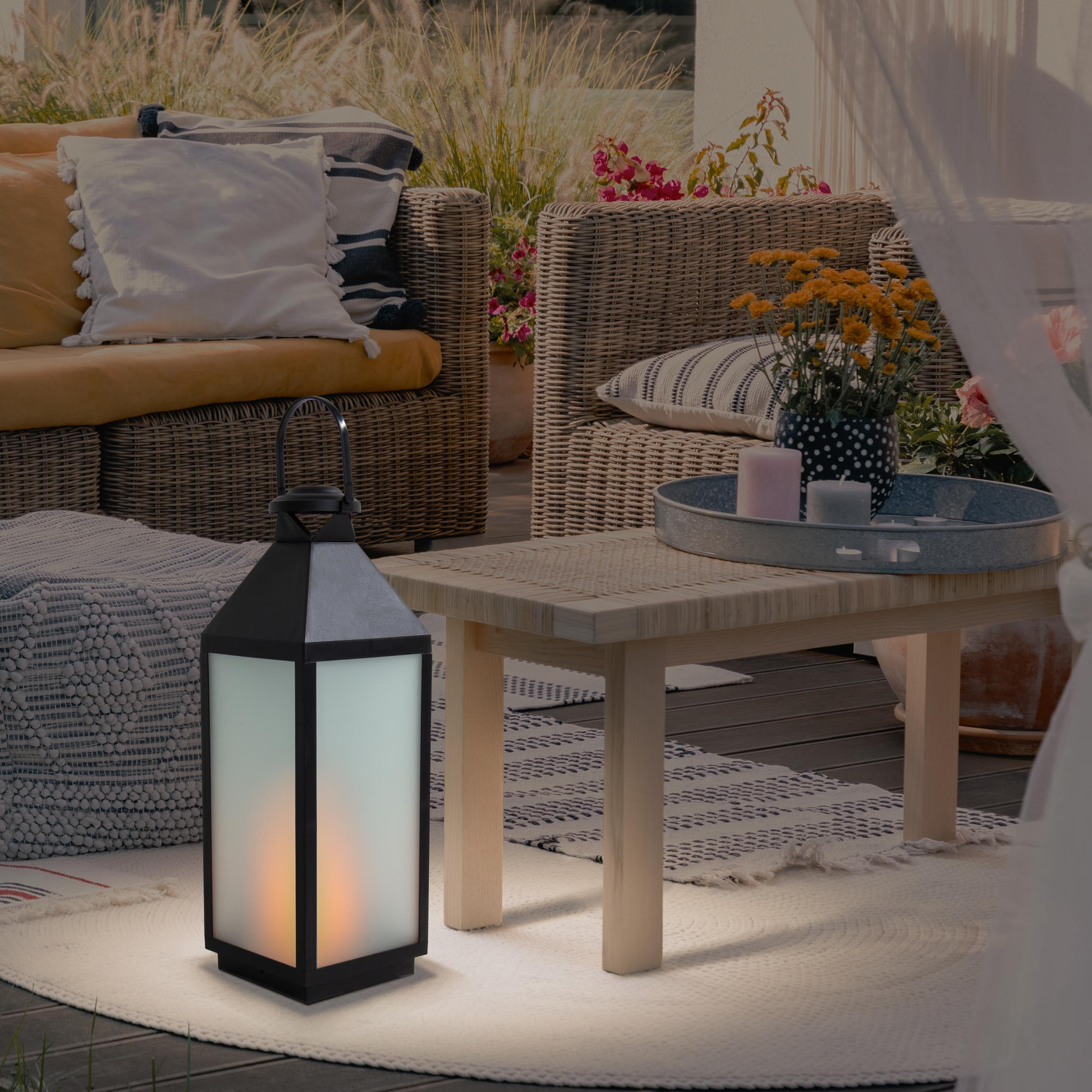 Eine schwarze Laterne auf einer Terrasse mit Tisch, Stühlen und Pflanzen. Die Laterne leuchtet mit einer Flammenattrappe.