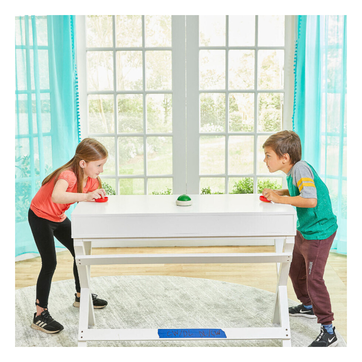 Dwoje dzieci grających w grę przy białym stole z zielonym przyciskiem. Zasłony i okno w tle.