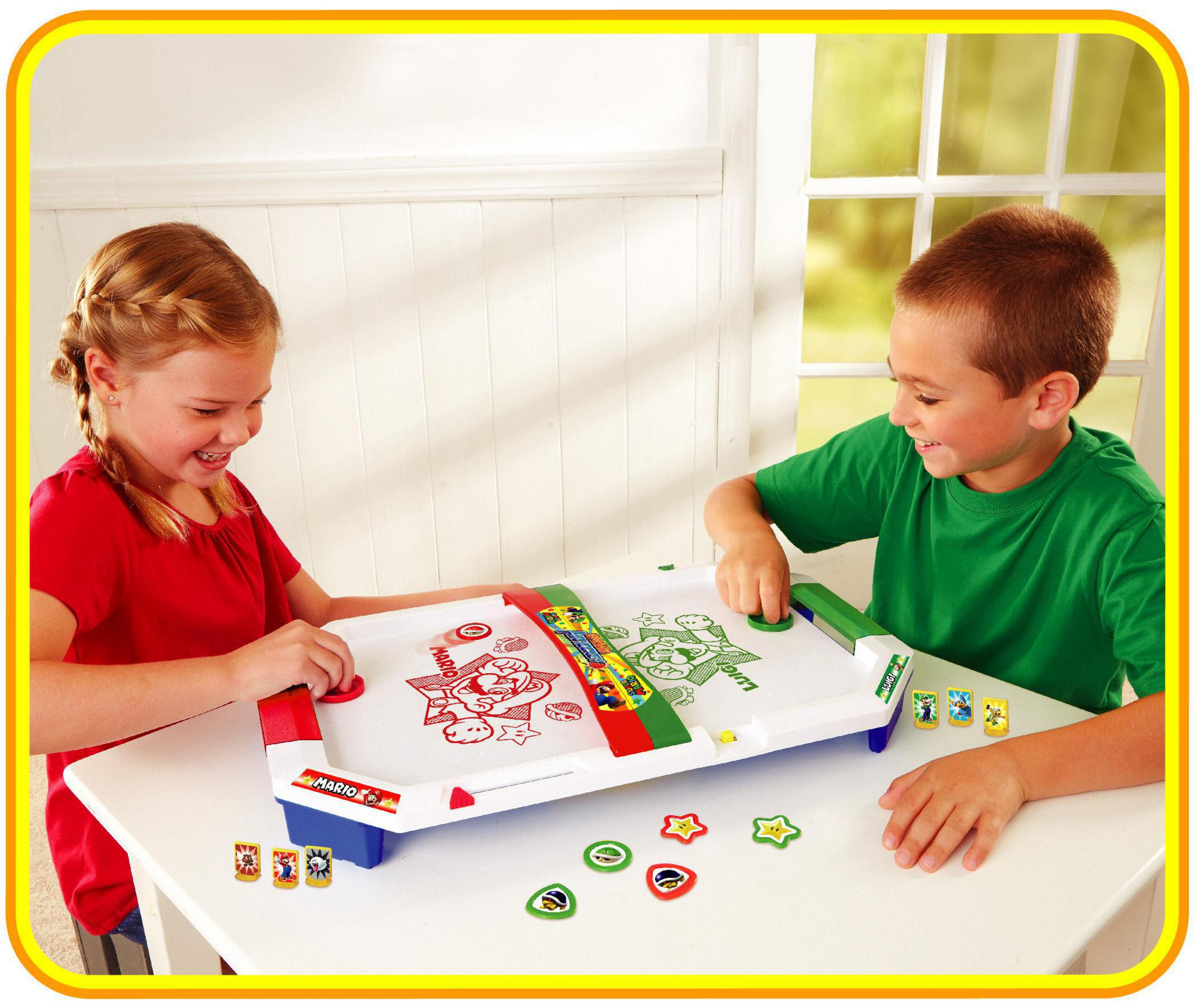 Two children play an air hockey game. The table is white with red, green, and cartoon designs.