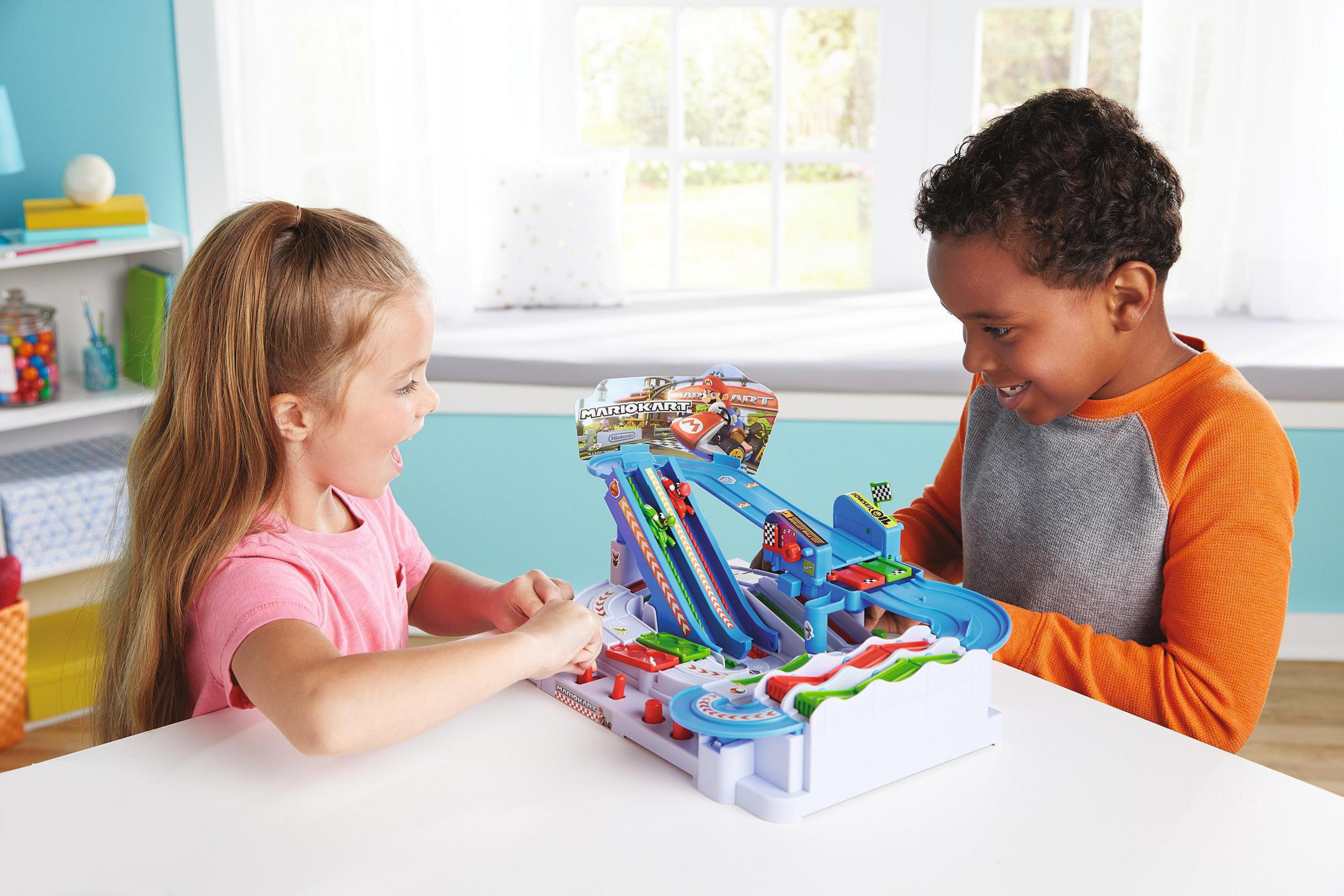 Dwoje dzieci bawiących się torem wyścigowym Mario Kart. Podekscytowane miny. Biały stół, niebieskie tło.