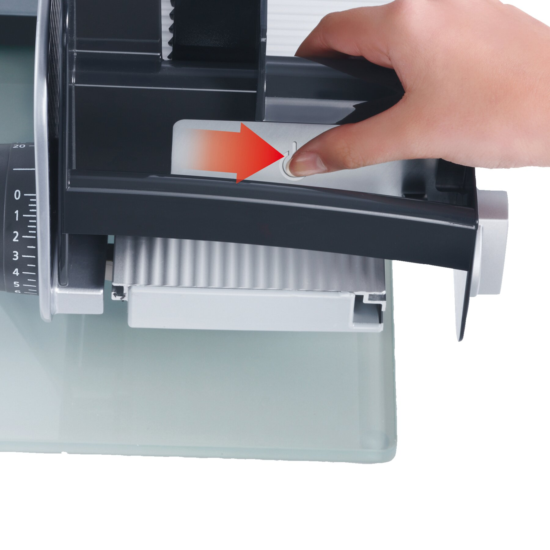 Ręka naciska biały przycisk na krajalnicy. Krajalnica jest srebrna i czarna. Poniżej szklana powierzchnia.