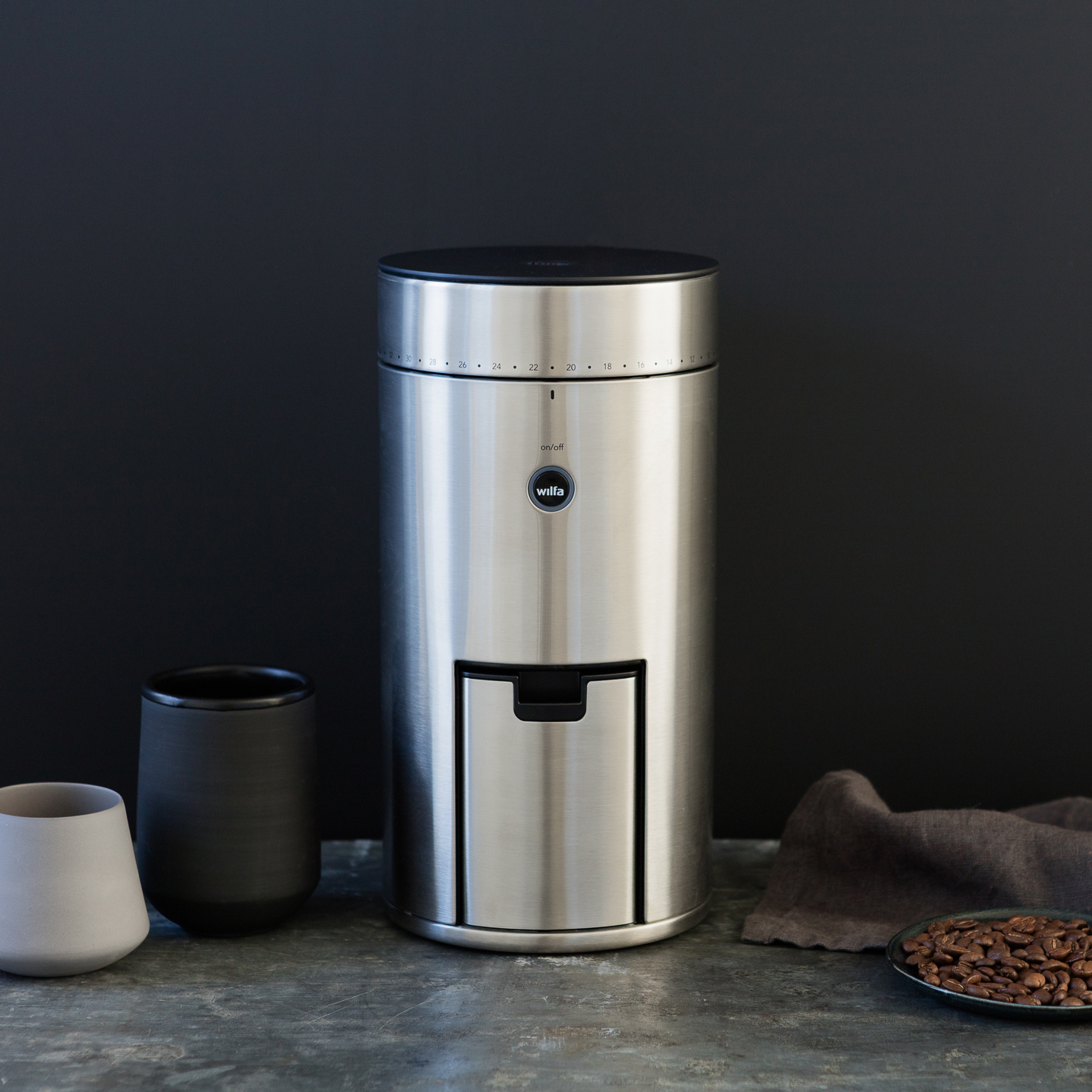 Eine Kaffeemühle aus Edelstahl mit schwarzem Deckel und Schublade. Hintergrund: Tassen und Bohnen.