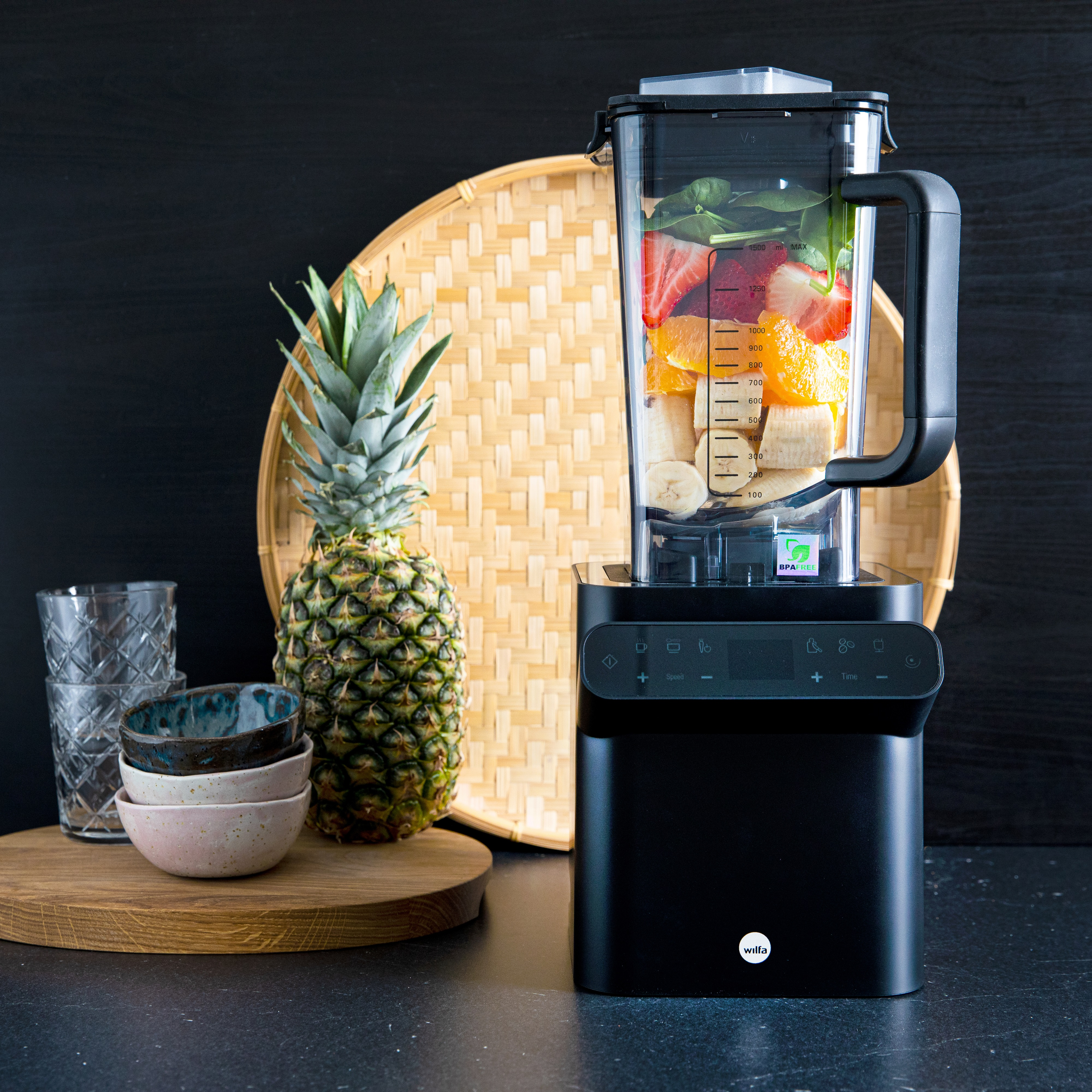 Mixer gefüllt mit Früchten auf schwarzer Theke. Ananas und Schüsseln daneben.