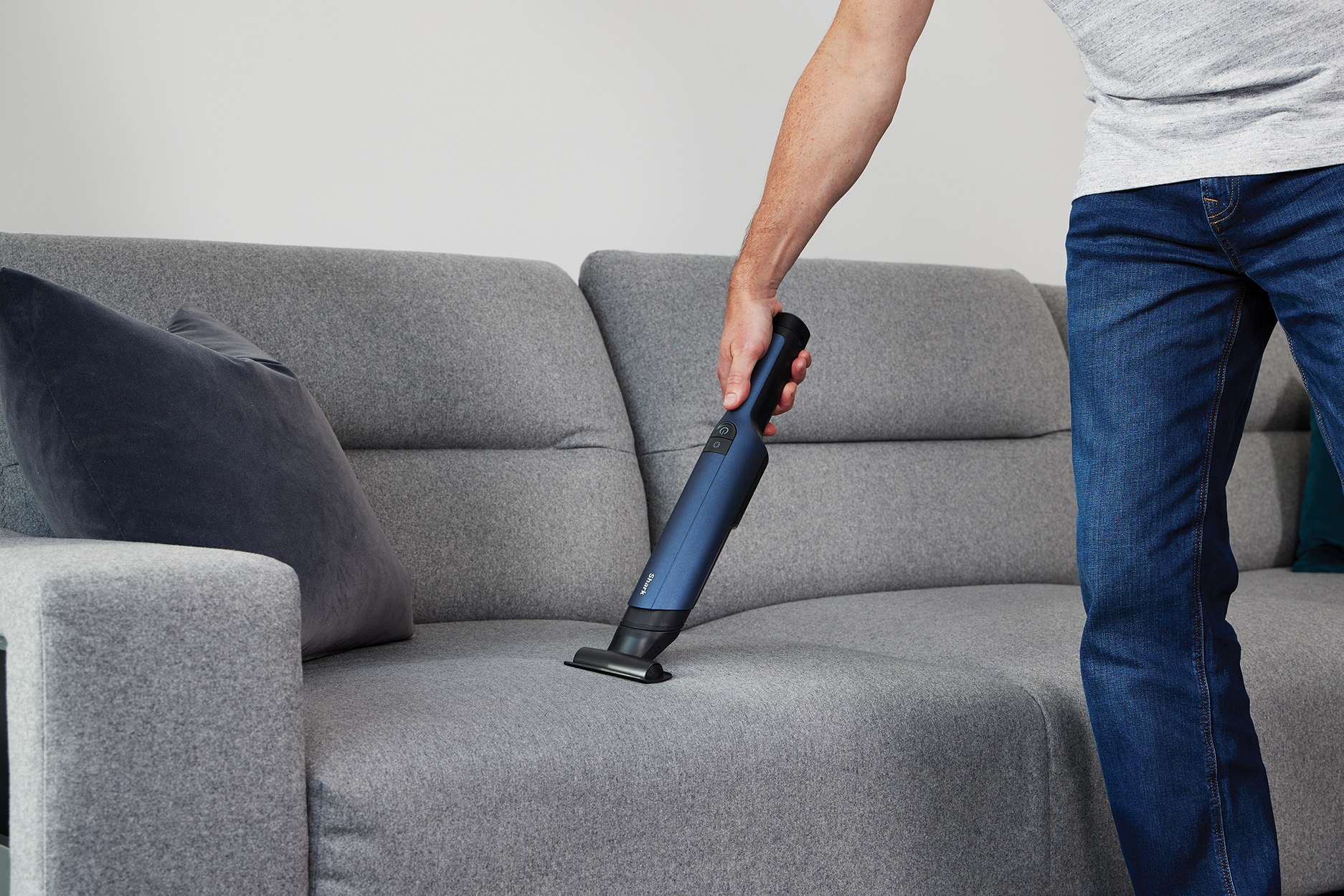 Mężczyzna odkurza szarą sofę niebiesko-czarnym odkurzaczem ręcznym.