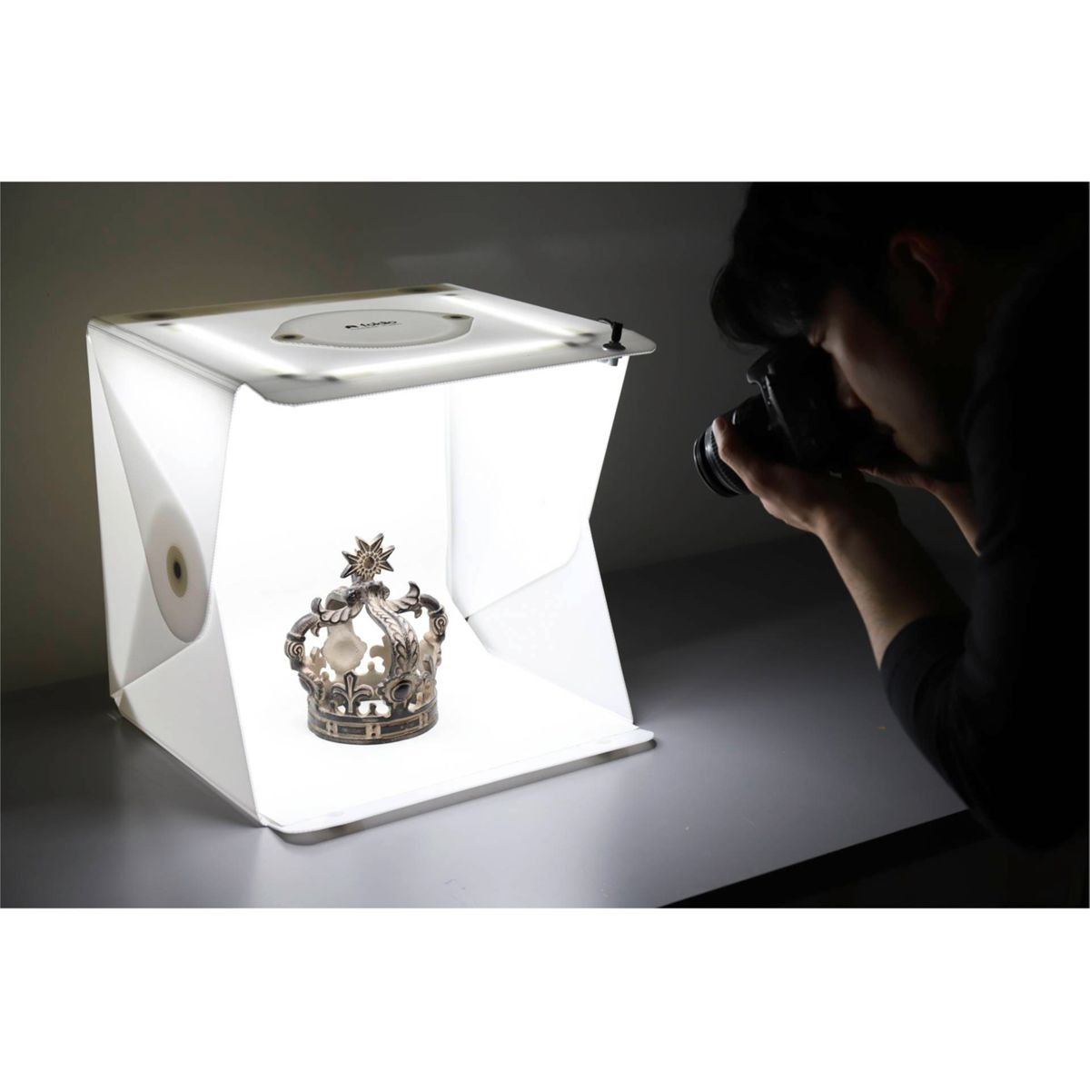 Eine Person fotografiert eine Krone in einer weißen Fotobox. Die Box ist beleuchtet, und die Krone liegt darin.