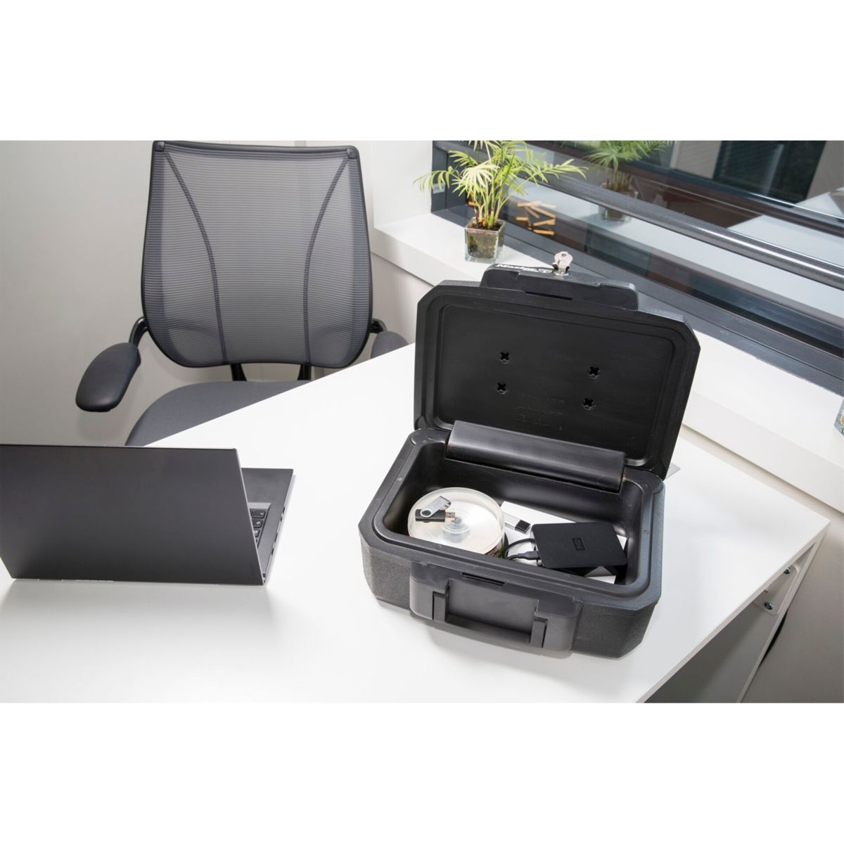 Ein schwarzer Safe, der auf einem weißen Schreibtisch geöffnet ist. Im Inneren befinden sich Gegenstände wie ein USB-Stick und eine Lupe.