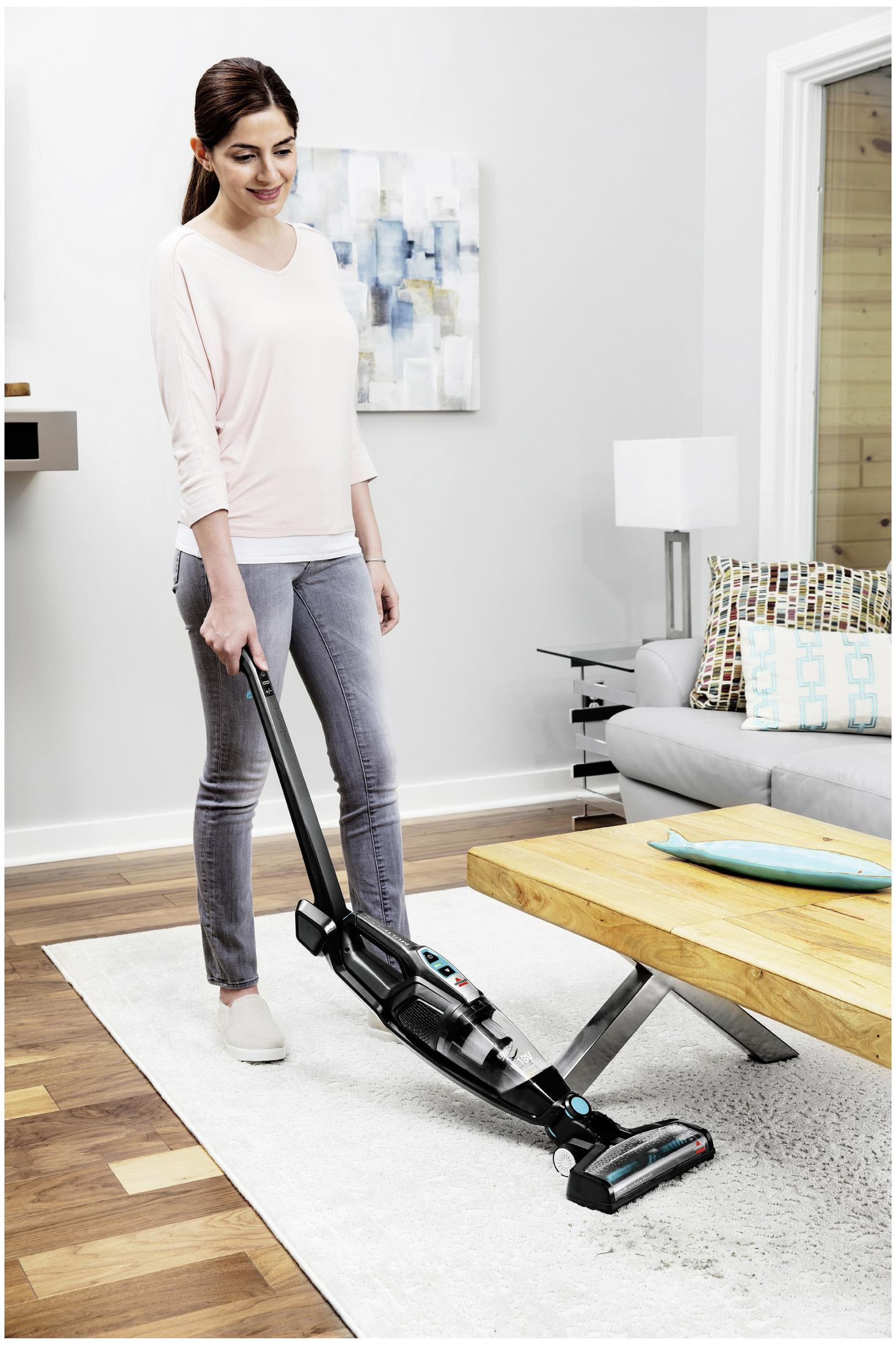 Kobieta odkurza dywan w salonie, trzymając odkurzacz pionowy.