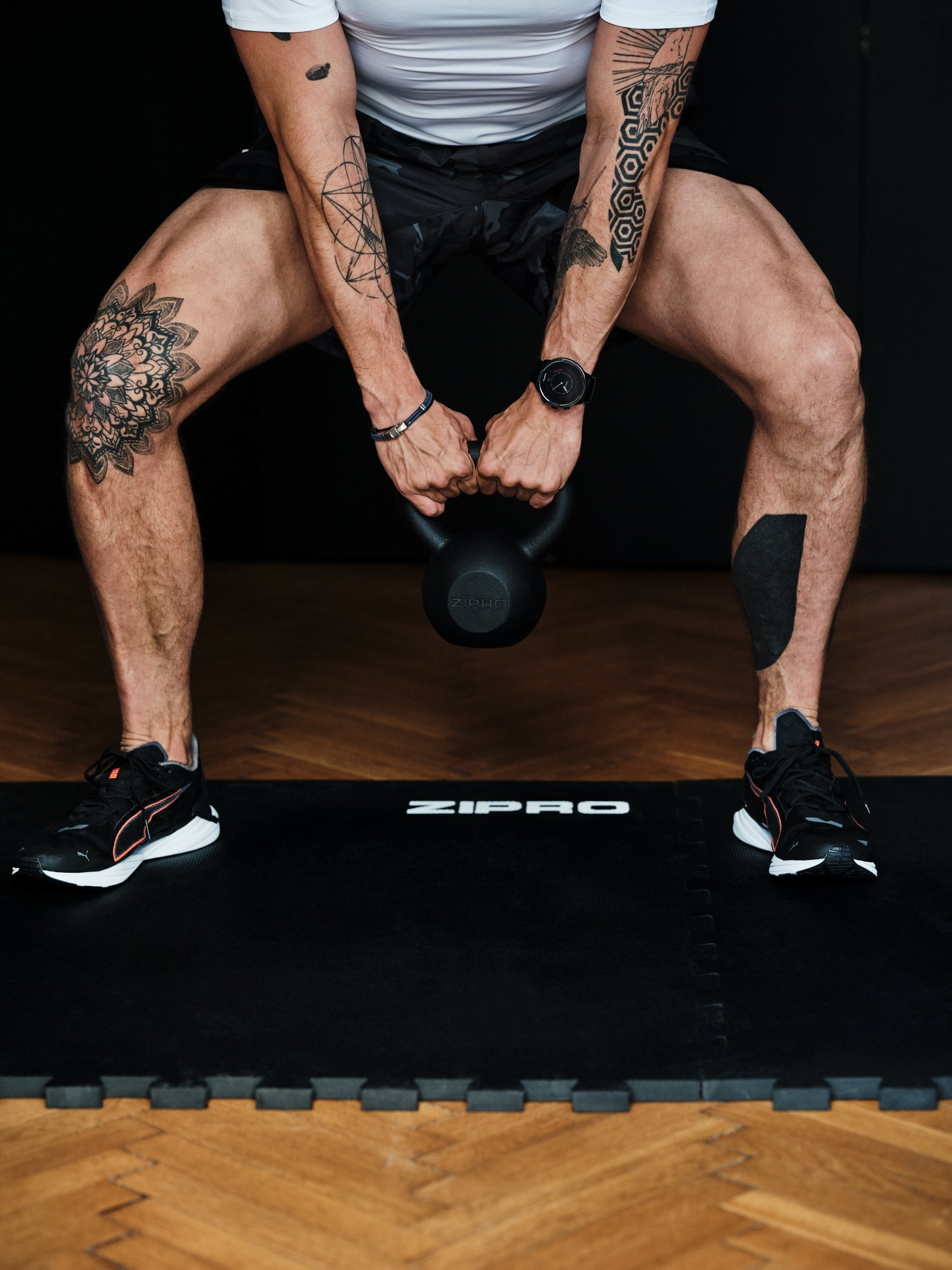 Ein Mann hält eine Kettlebell in der Hocke. Tätowierungen auf Beinen und Armen sichtbar.