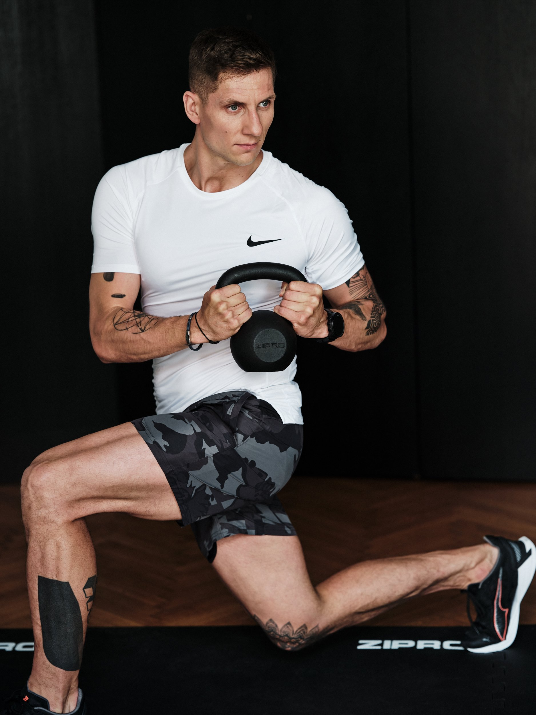 Mann macht eine Ausfallschrittübung, hält eine Kettlebell im Fitnessstudio. Er trägt ein weißes Hemd und Camouflage-Shorts.