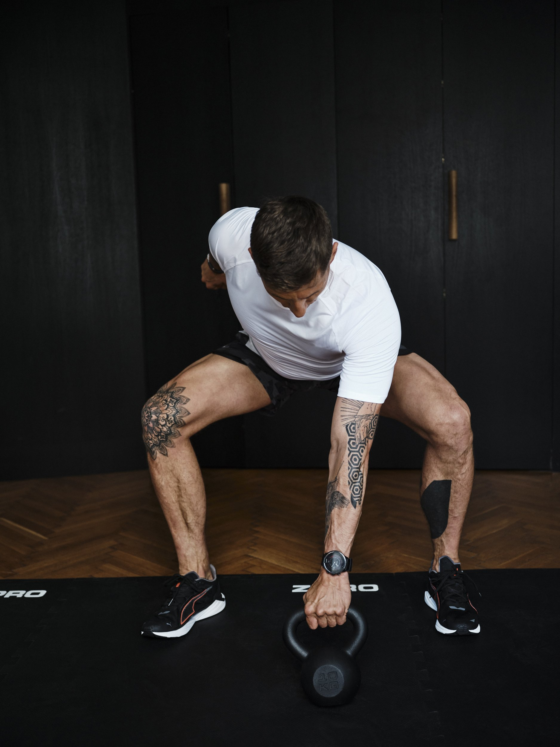 Ein Mann macht eine Kettlebell-Übung in einem Fitnessstudio. Er trägt ein weißes Hemd und schwarze Shorts. Tätowierungen sind sichtbar.
