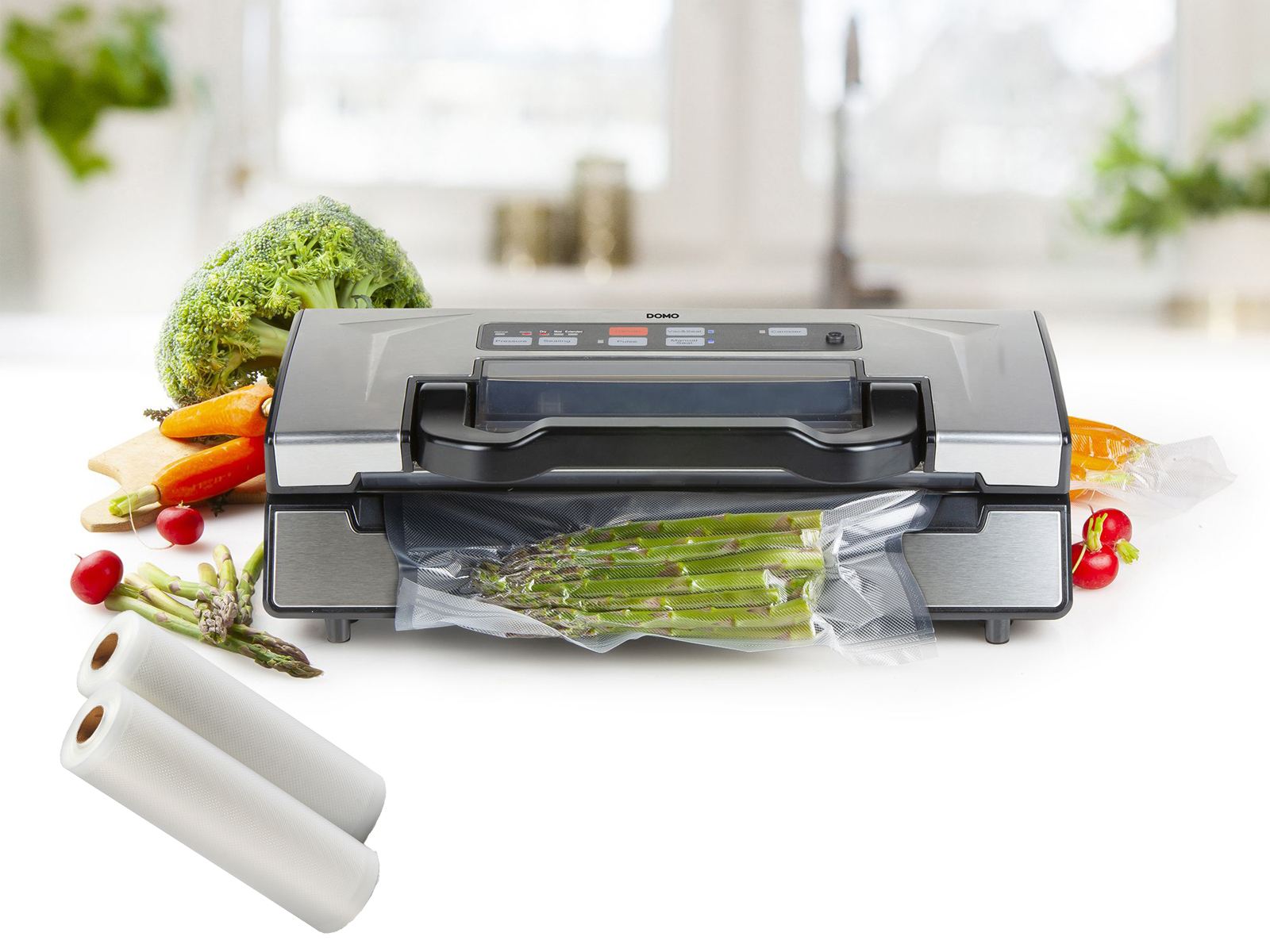 Silberner Vakuumierer mit Spargel in einer Tüte, umgeben von Gemüse und Plastikrollen.