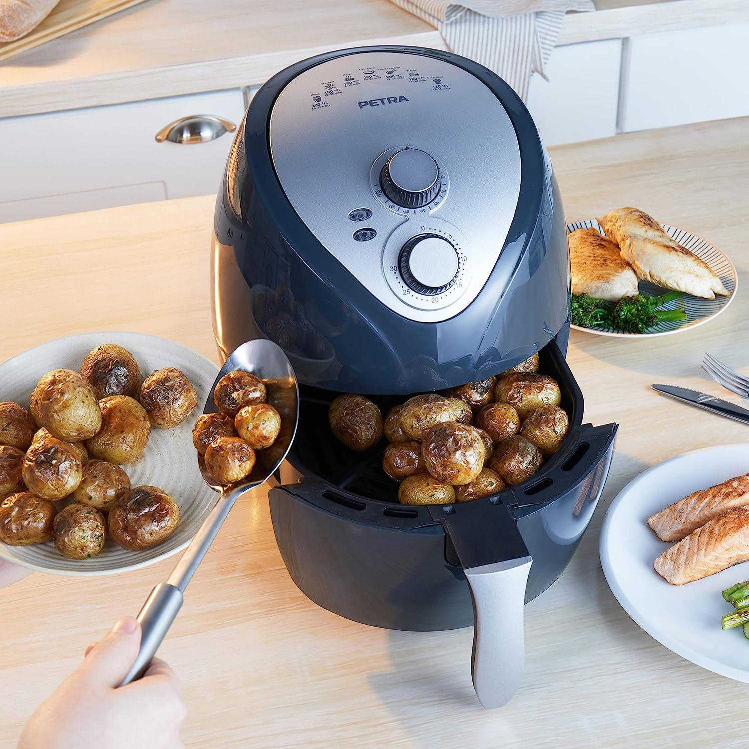 Eine Heißluftfritteuse mit Kartoffeln, die herausgeschöpft werden. Gekochtes Huhn und Lachs auf Tellern daneben.