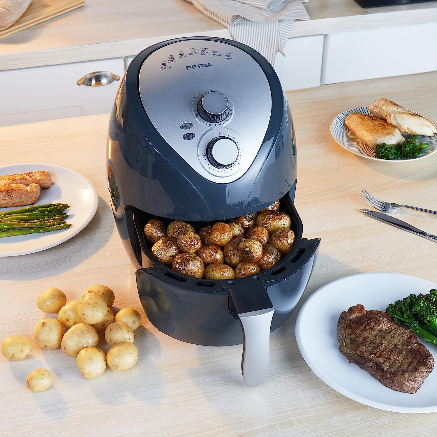Eine Heißluftfritteuse mit Kartoffeln und Essen. Gemüse und Steak sind auf Tellern. Küchenszene.
