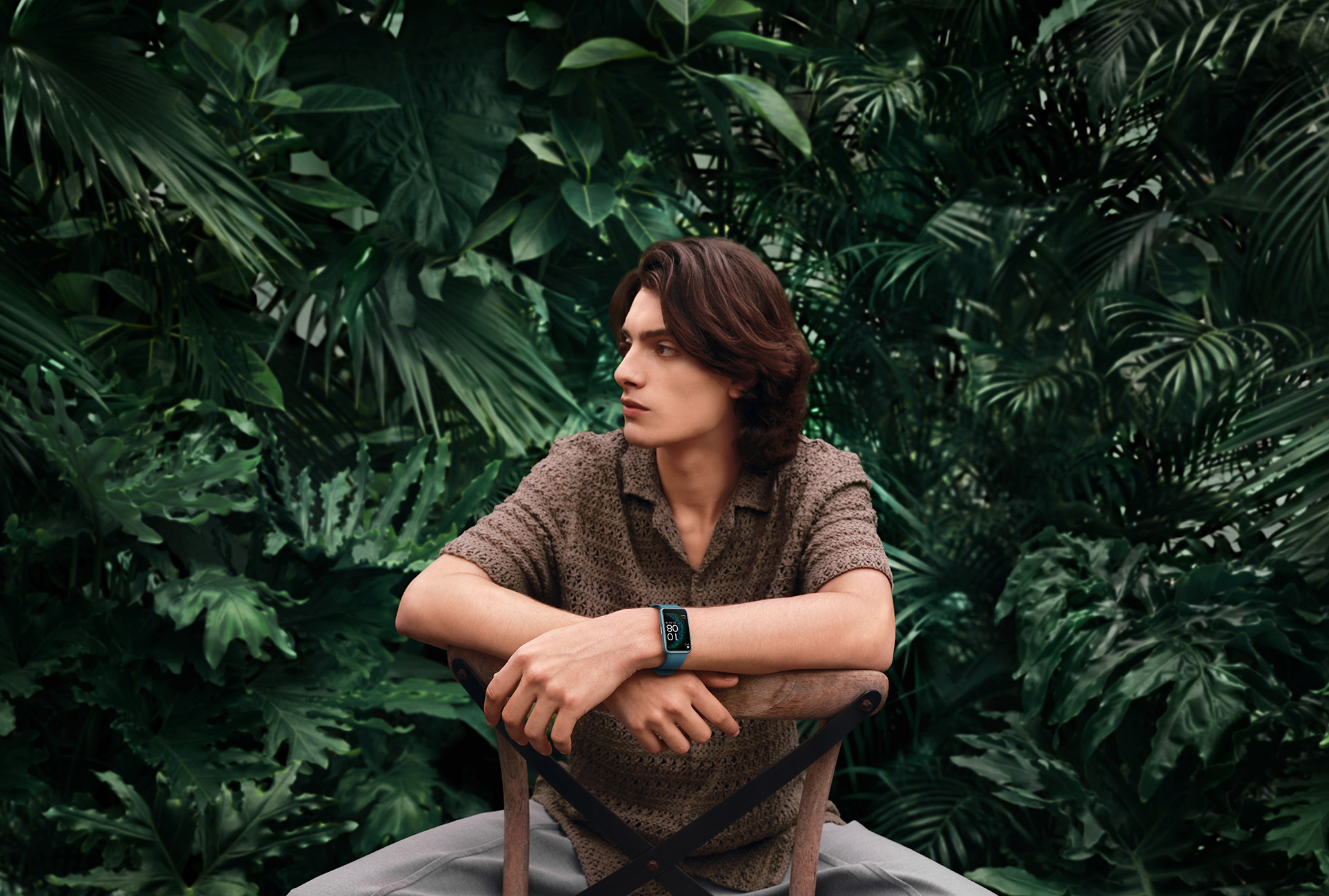 Mężczyzna siedzi na drewnianym krześle, w koszuli i turkusowym smartwatchu, otoczony zielonymi liśćmi.