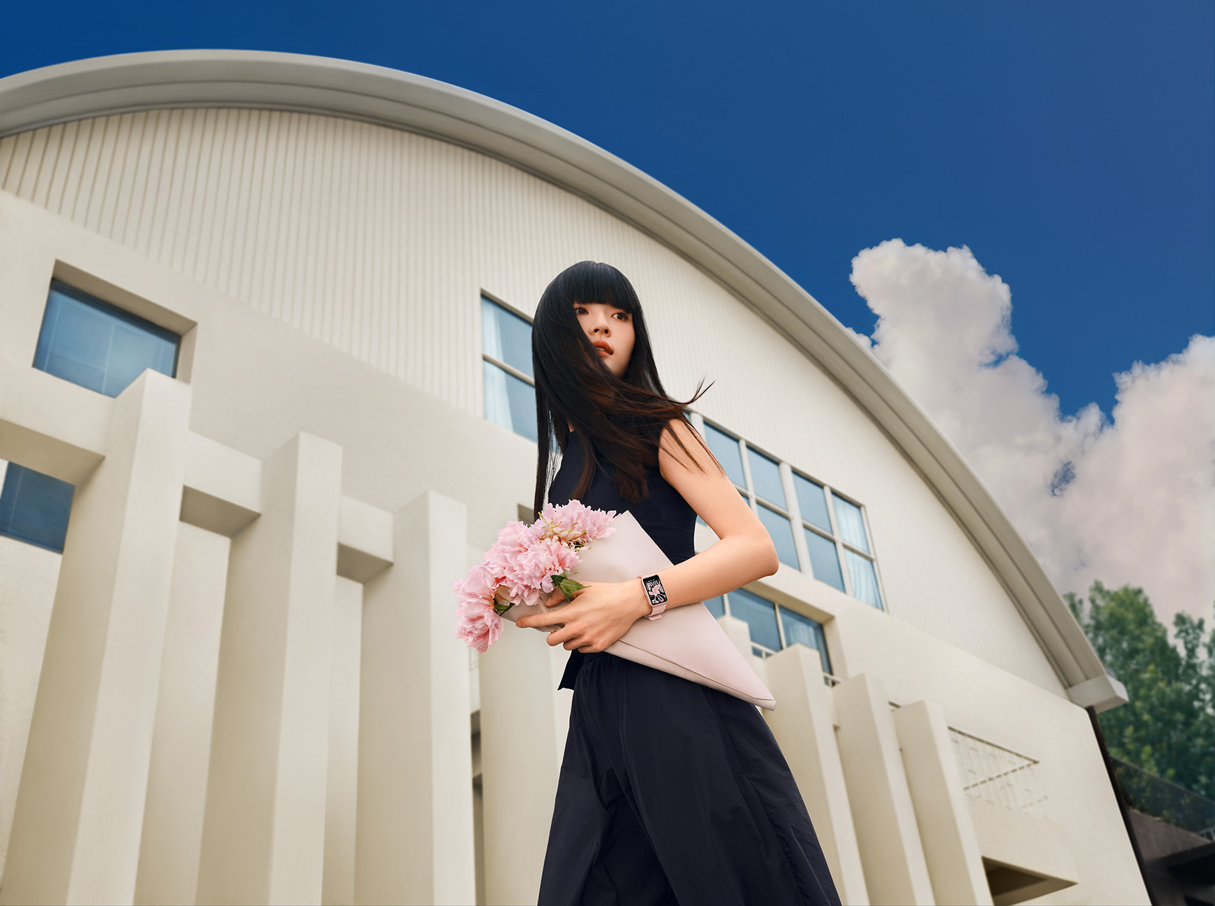 Vrouw in zwarte kleding met bloemen voor een wit gebouw onder een blauwe hemel.
