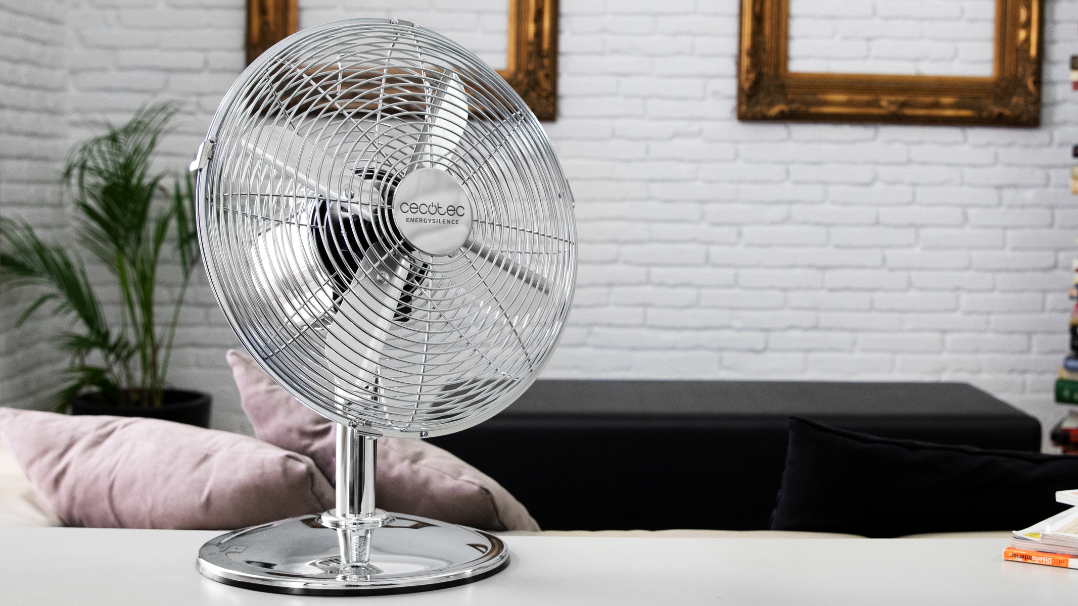 Ventilatore argentato su un tavolo con oggetti decorativi sullo sfondo.