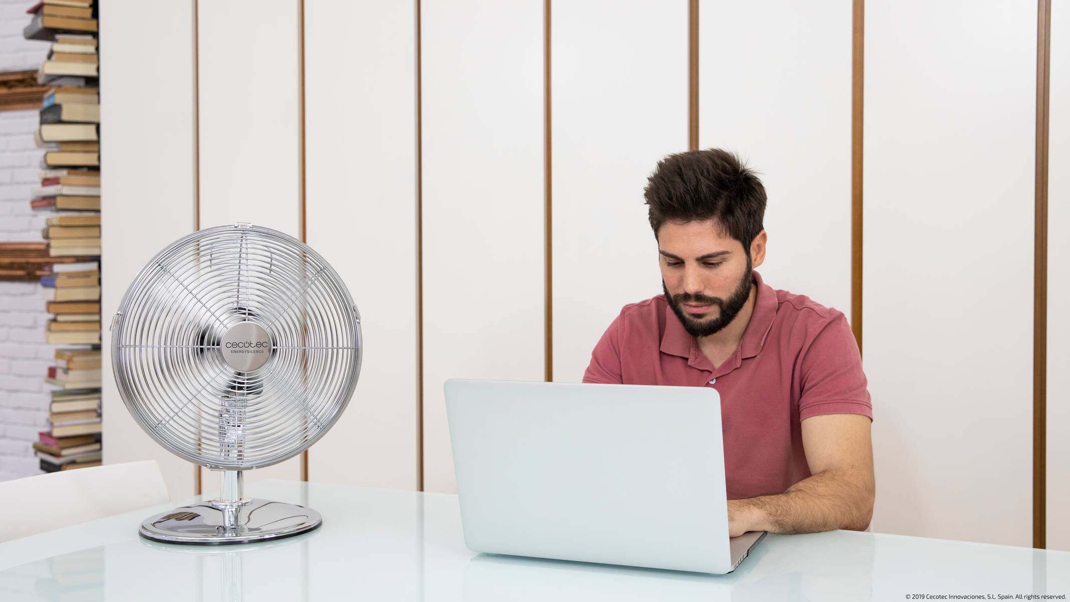Un uomo usa un laptop seduto a un tavolo con un ventilatore argentato. Librerie sullo sfondo.