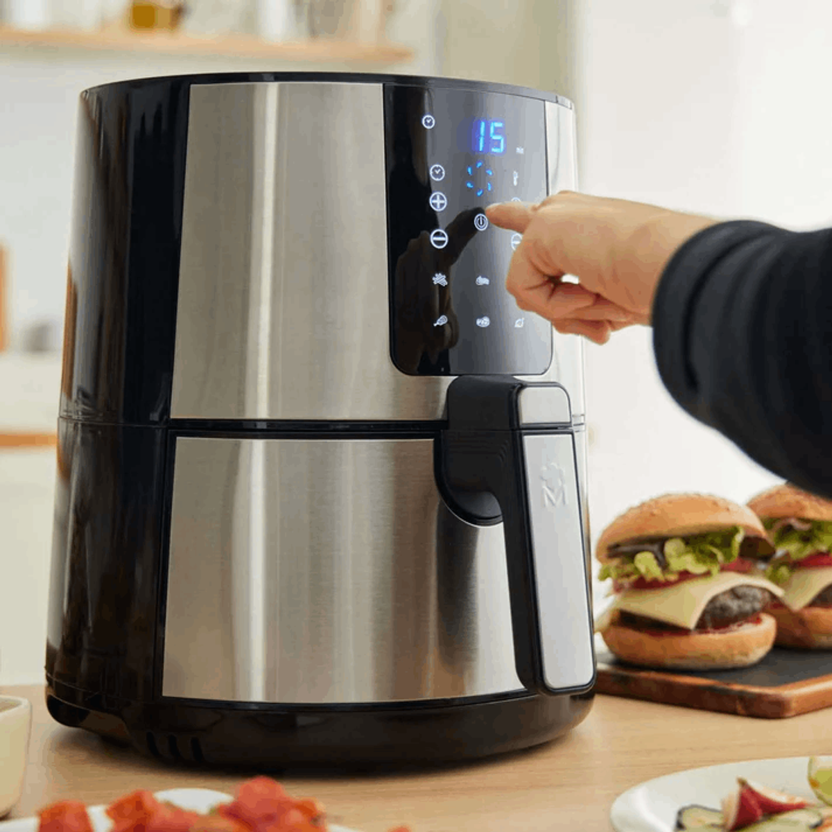 Una friggitrice ad aria con mano che tocca il display, e hamburger sullo sfondo.