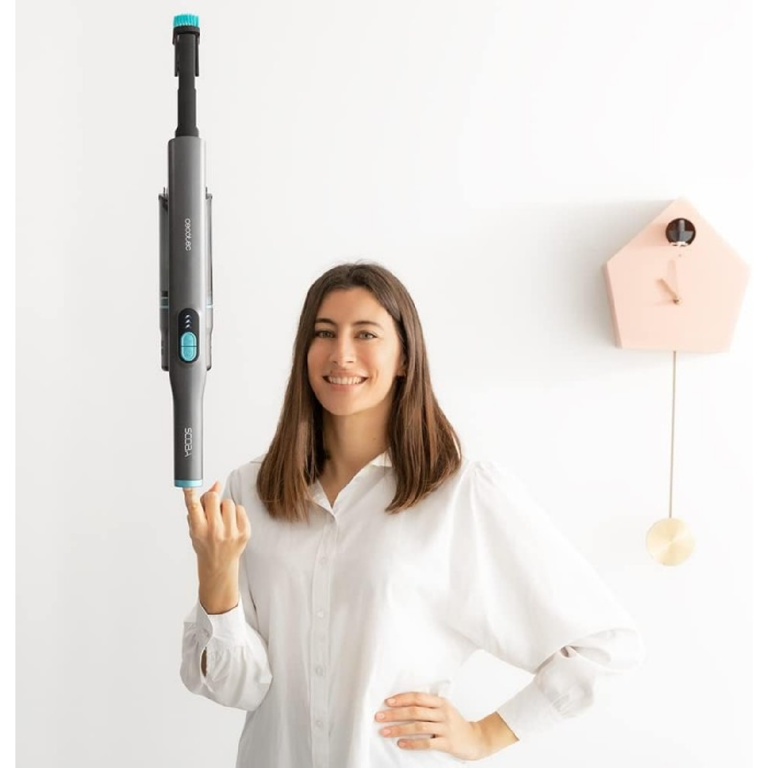 Mujer sosteniendo una aspiradora inalámbrica. Sonriendo, pared blanca y reloj rosa.