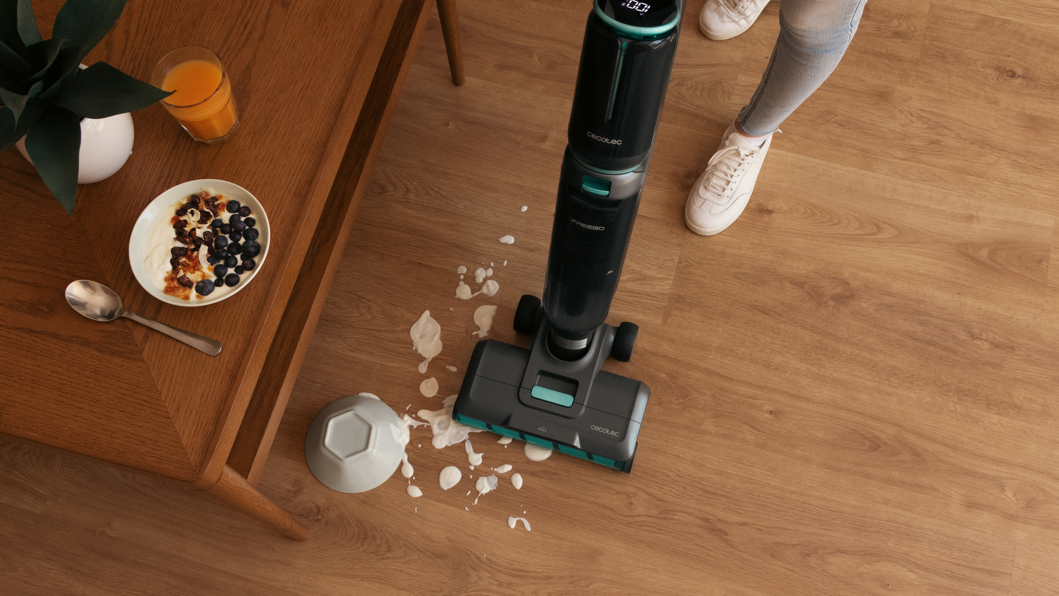 Un aspirapolvere per pavimenti che aspira un pavimento in legno. Sono visibili una ciotola di latte e cibo versati. C'è una persona nell'immagine.