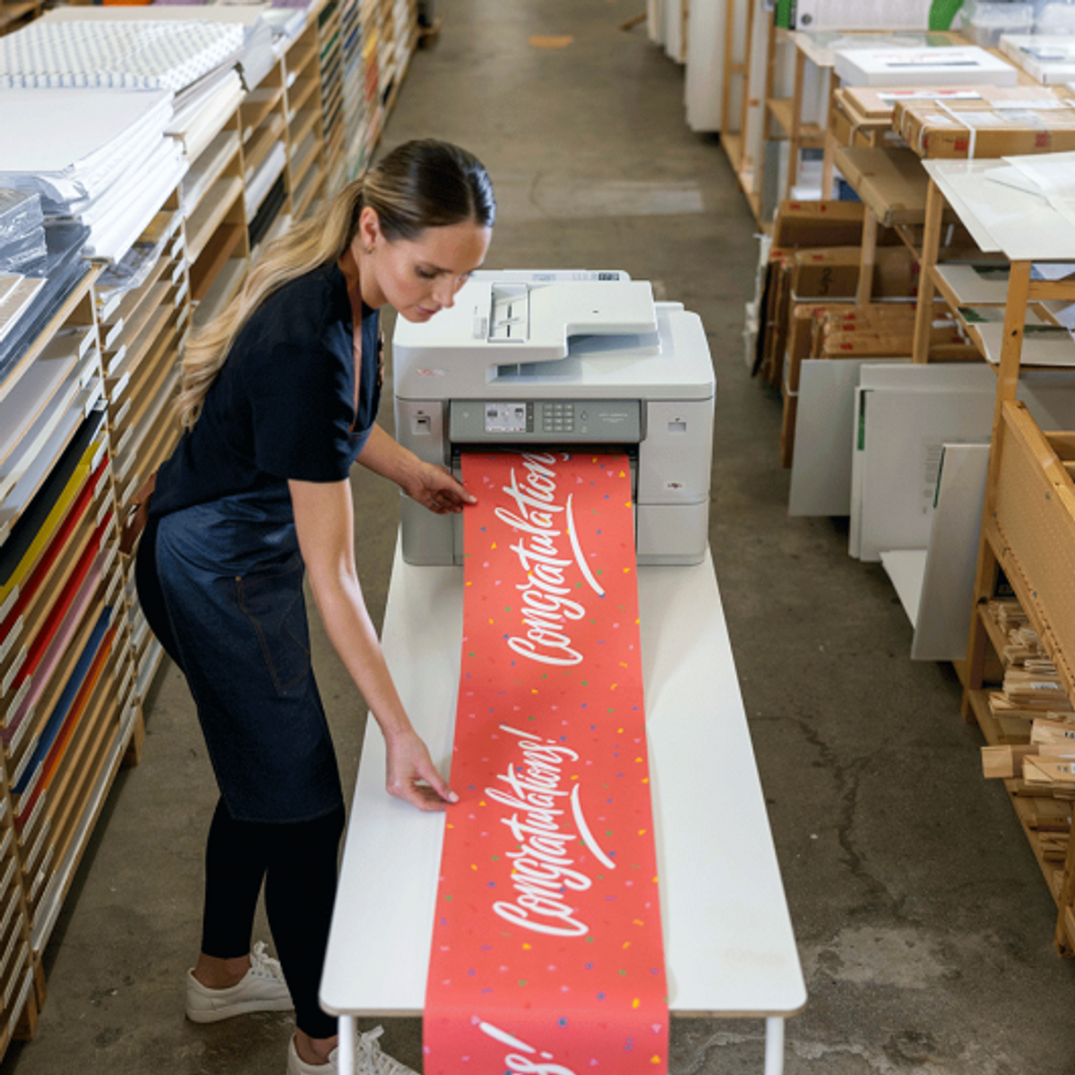 Frau druckt rotes Banner mit "Herzlichen Glückwunsch!" auf großem Drucker.