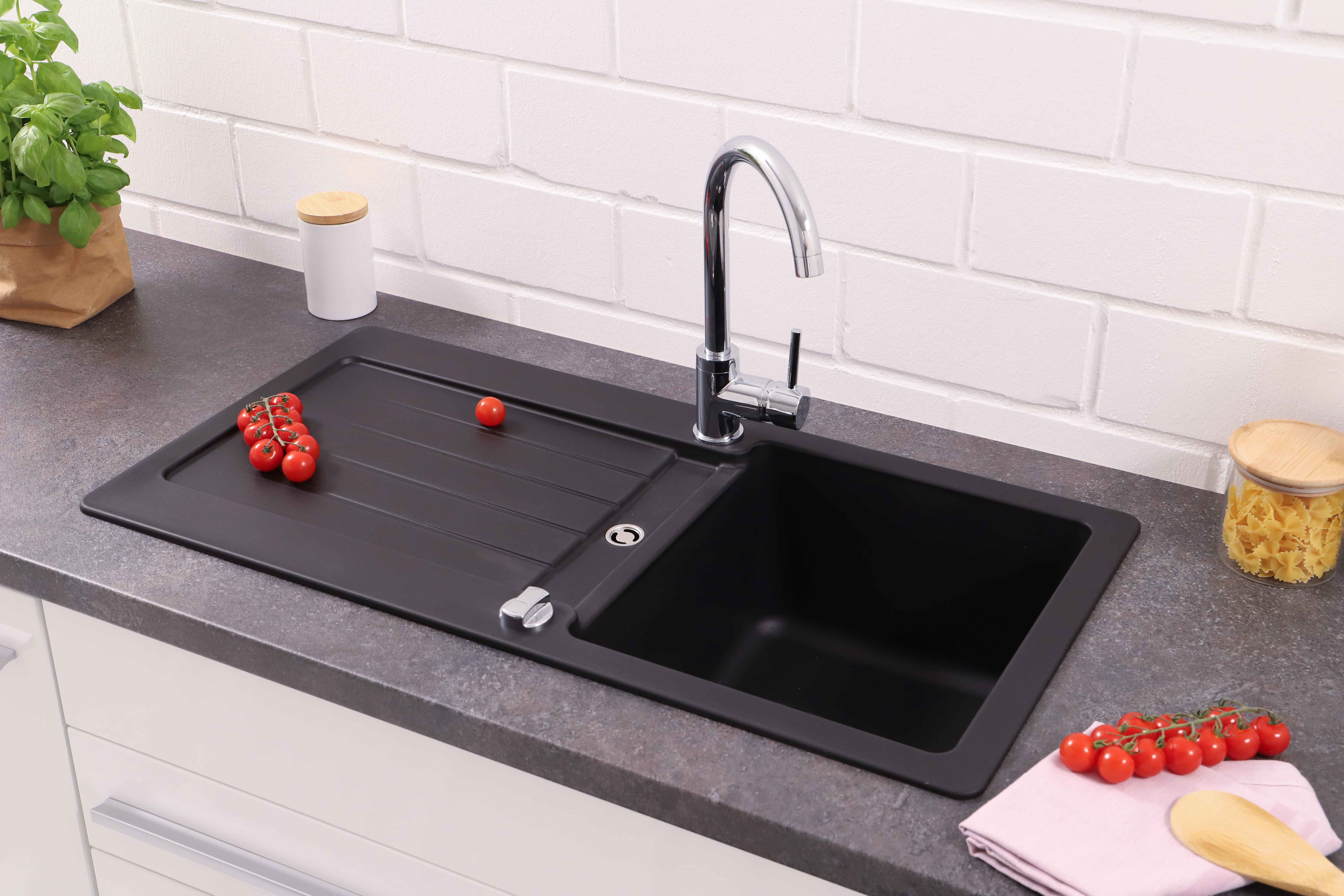 Schwarzes Küchenbecken mit Chromarmatur. Tomaten und Pasta liegen auf der Arbeitsplatte. Weißer Ziegelhintergrund.