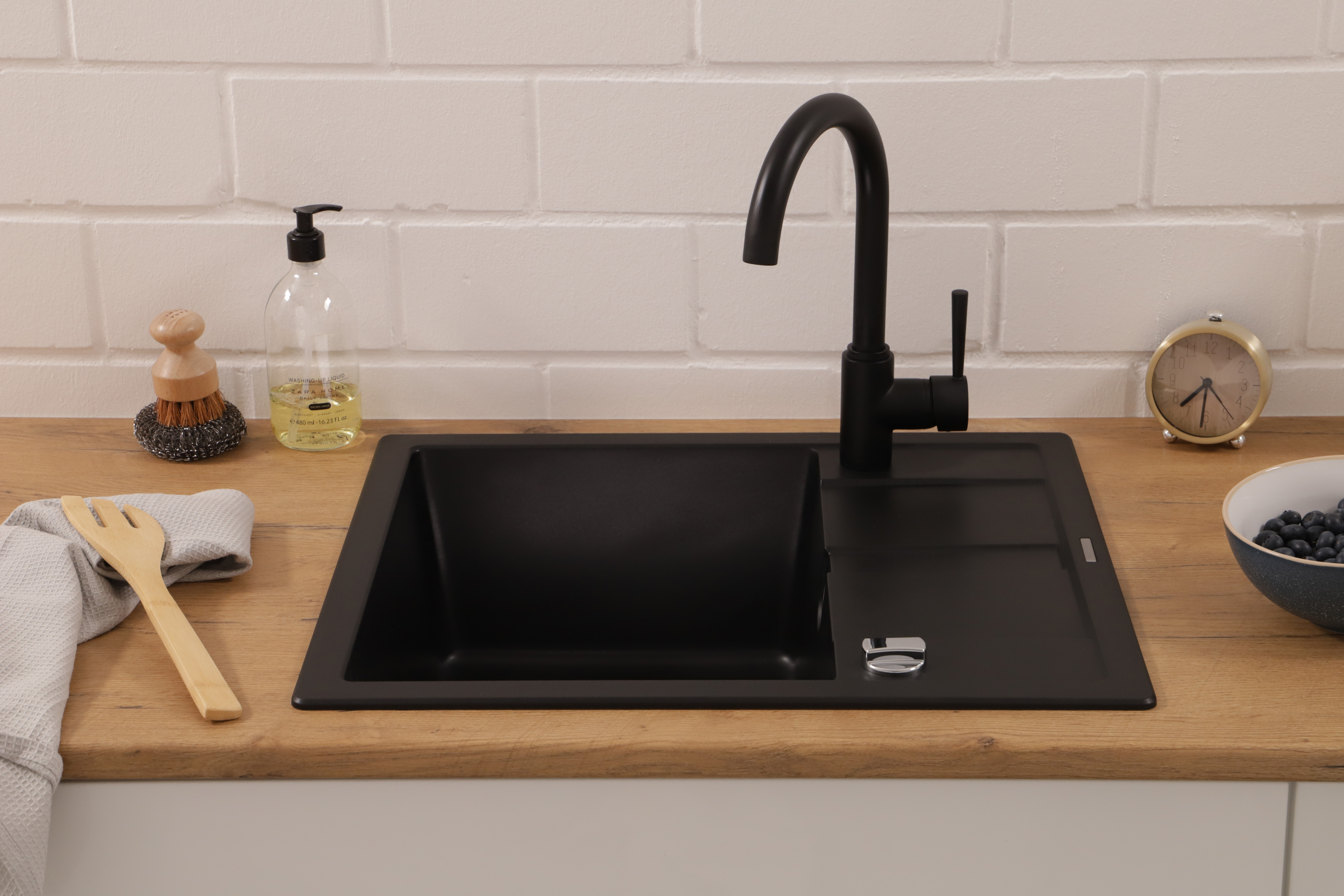 Lavello nero con rubinetto nero. Piano di lavoro in legno. Sono presenti sapone, spazzola, orologio e mirtilli.