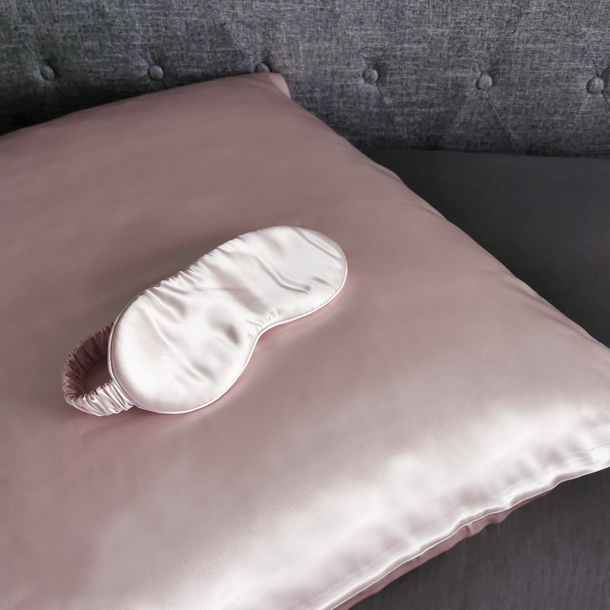 Rosa Satin-Kissen mit passender Schlafmaske auf einem grauen Bett.