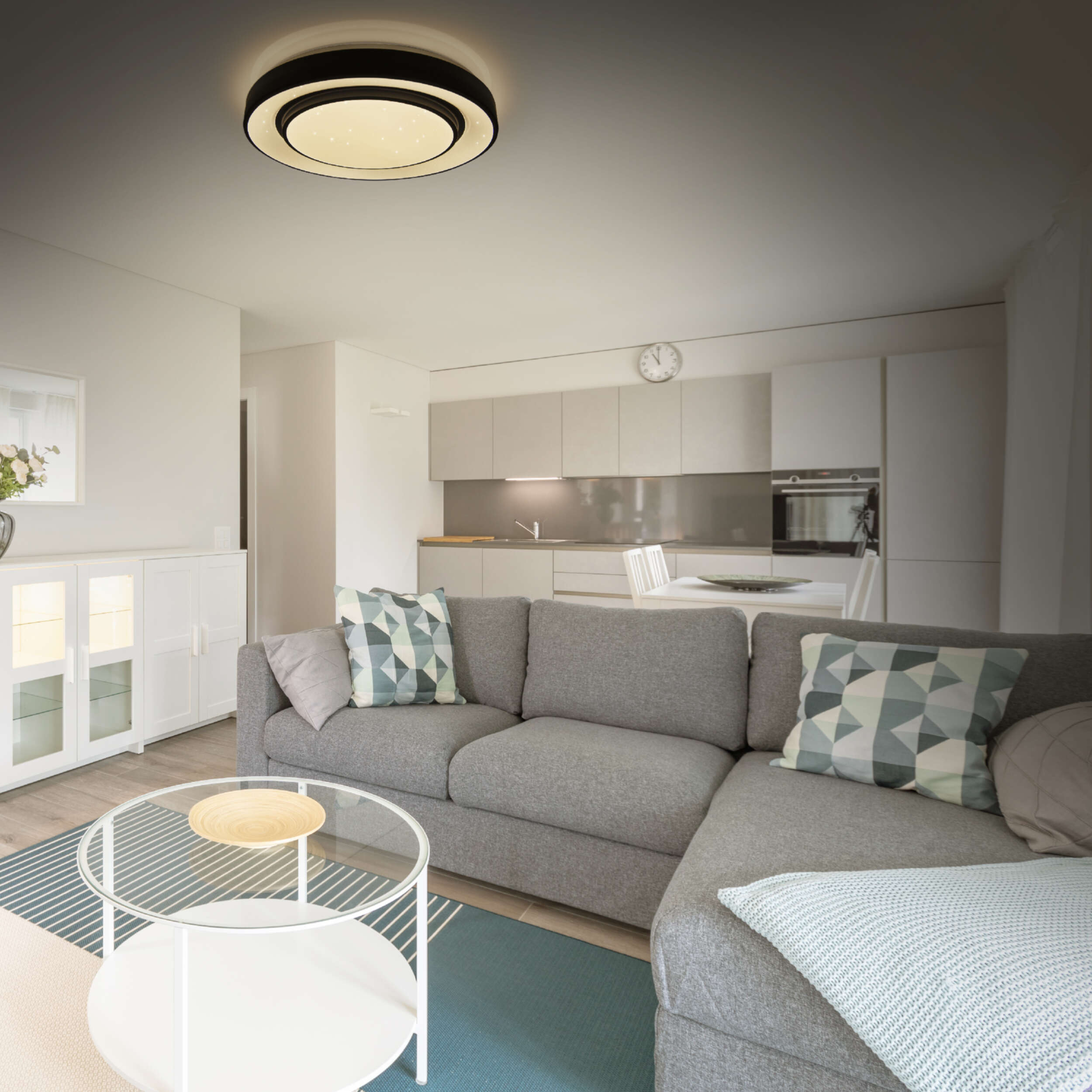 Ein Wohnzimmer mit grauem Sofa, Couchtisch und Deckenleuchte. Die Küche ist im Hintergrund.
