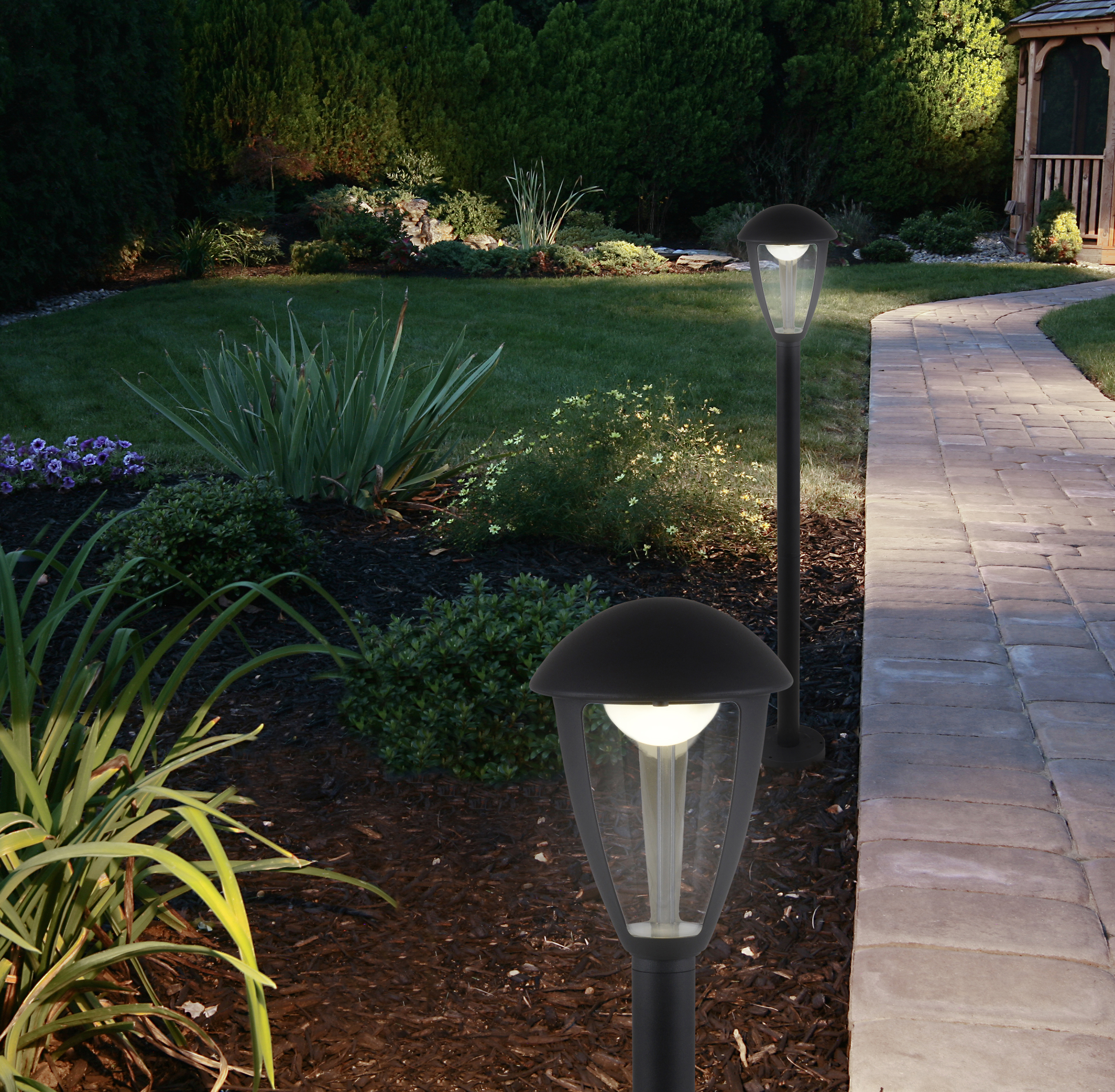 Zwei schwarze Außenlampen beleuchten nachts einen Gartenweg. Grünes Gebüsch und Gras umgeben den Weg.