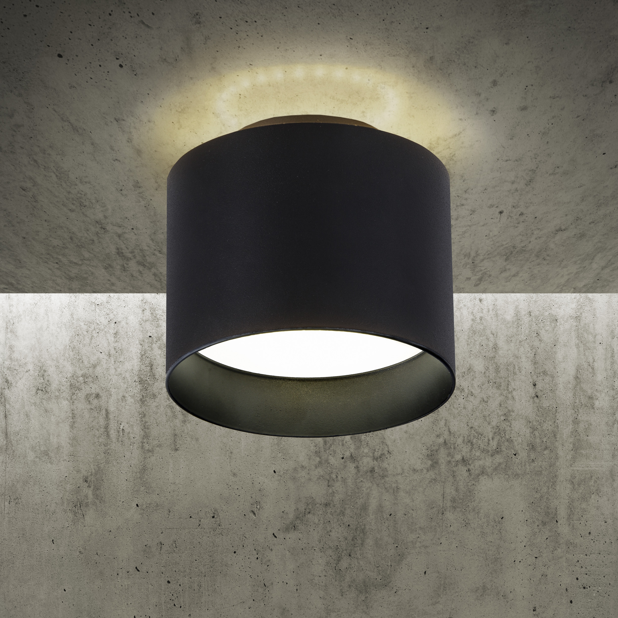 Eine schwarze runde Deckenlampe mit dunkelgrauem Schirm vor einer Betonwand.