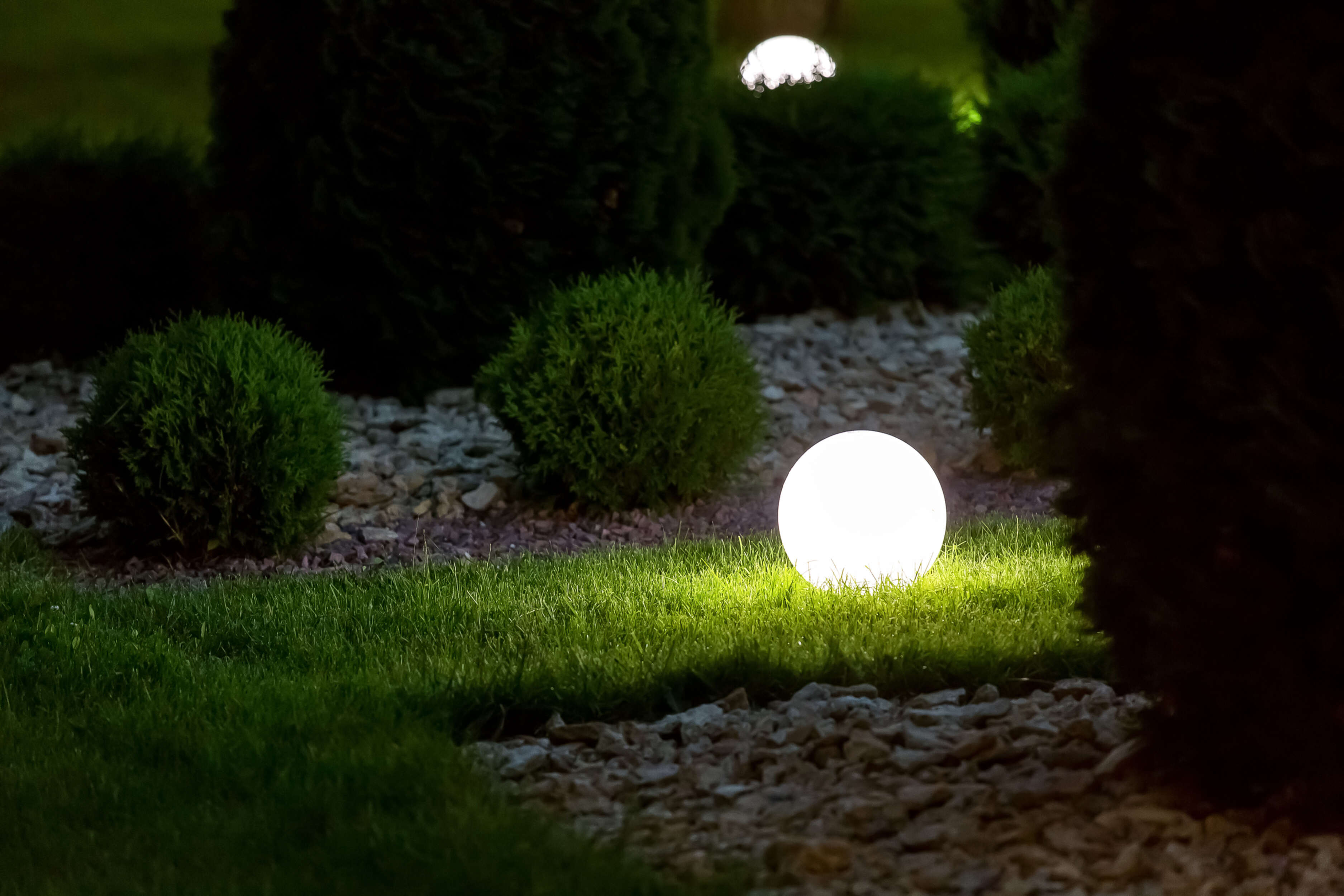 Gartenszene bei Nacht mit leuchtenden Kugellichtern auf Gras, umgeben von Büschen.